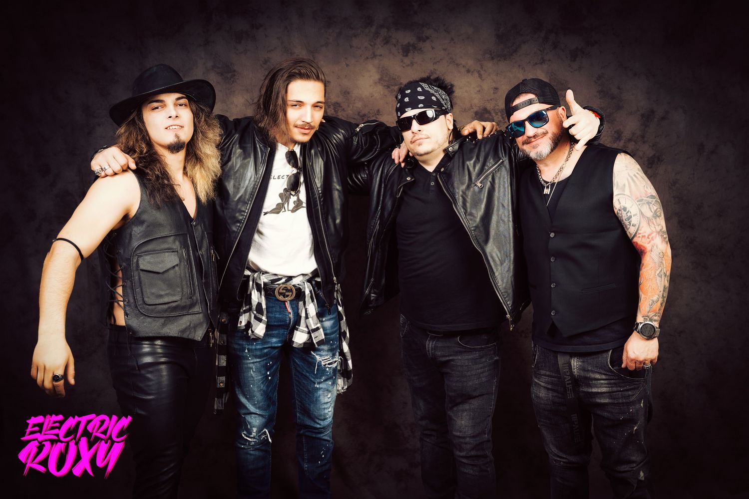 Four members of the rock band Electric Roxy in leather vests and jackets pose arm in arm against a dark textured studio backdrop.