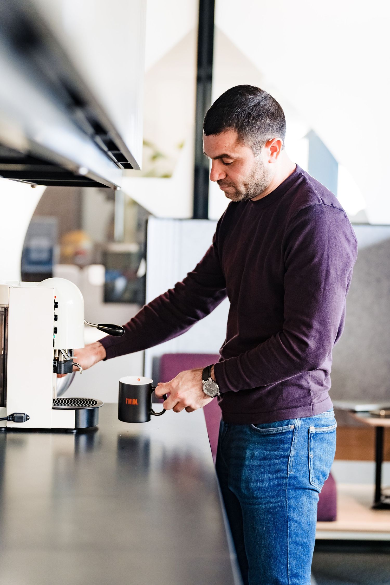 Man in a maroon sweater prepares coffee at an espresso machine while holding a black mug in a modern office kitchen.
