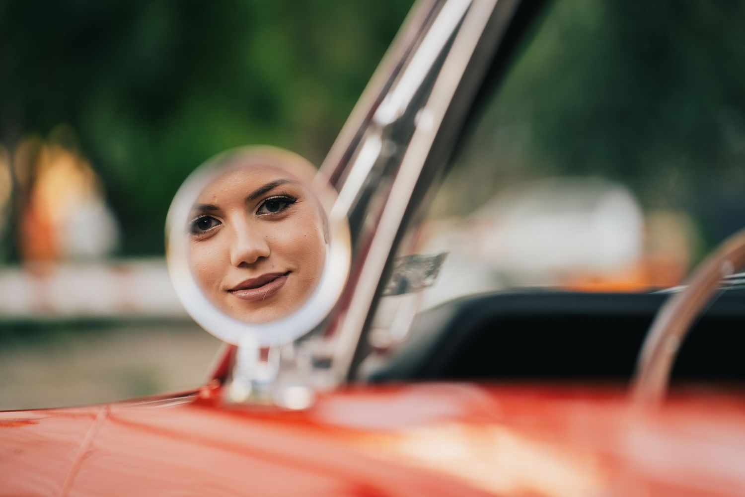 A smiling woman's face is reflected in the round side mirror of a red classic car, with a softly blurred green background.