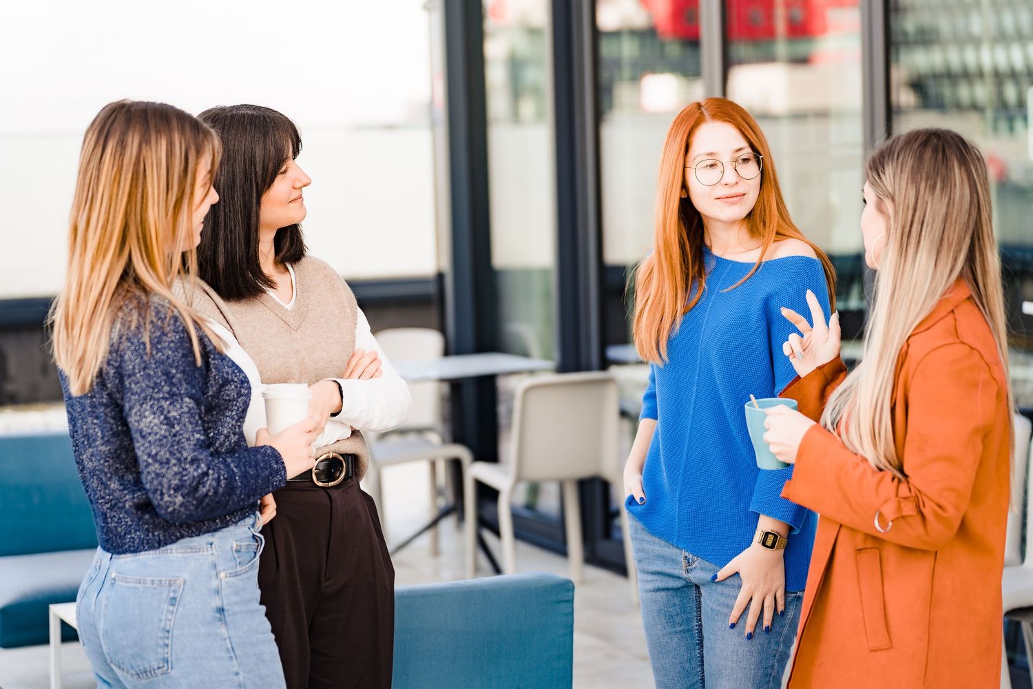Four women talk and laugh in a bright modern office lounge with glass windows and comfortable blue seating.