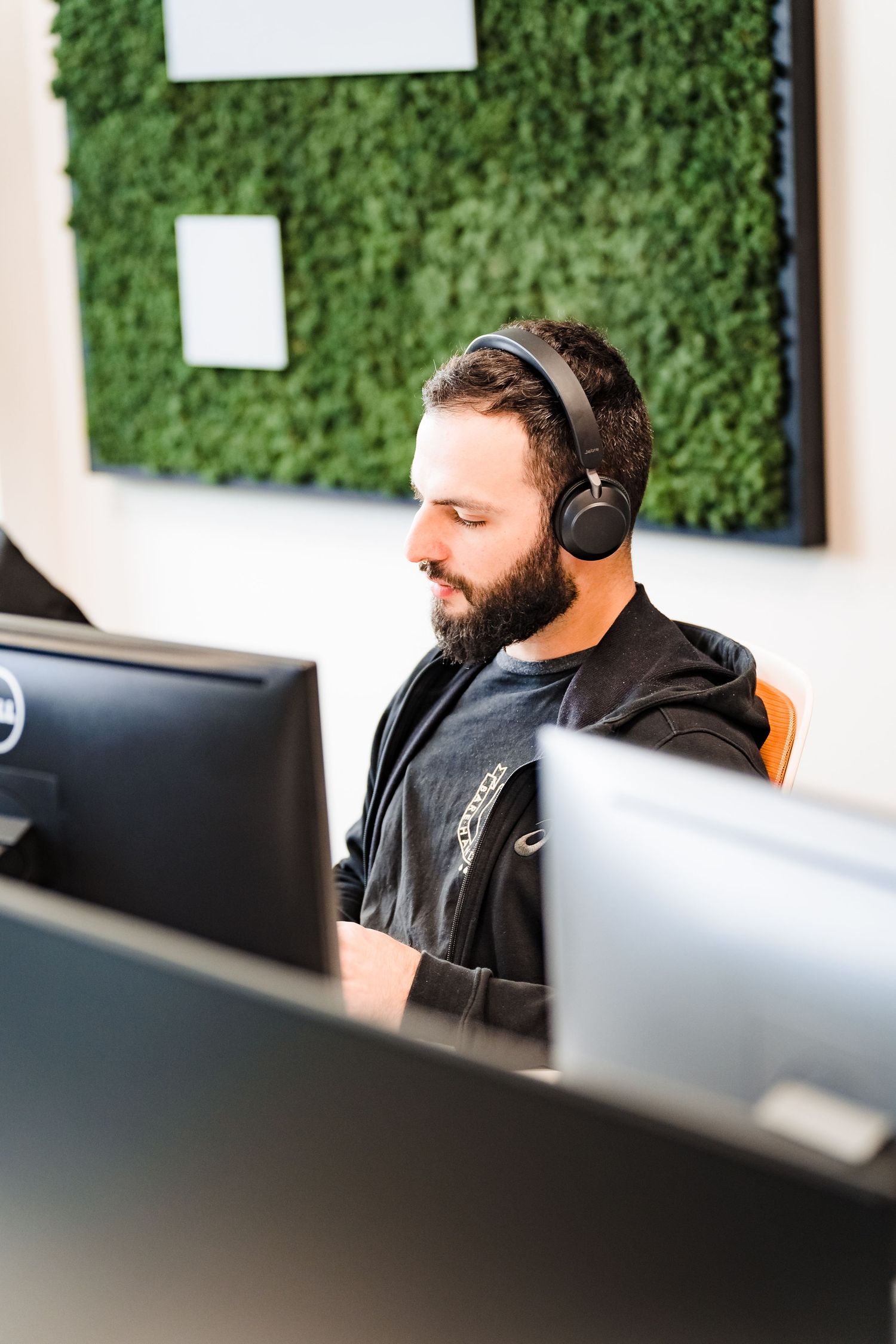 Bearded man wearing wireless headphones works at a computer desk in a modern office with a green moss wall behind him.