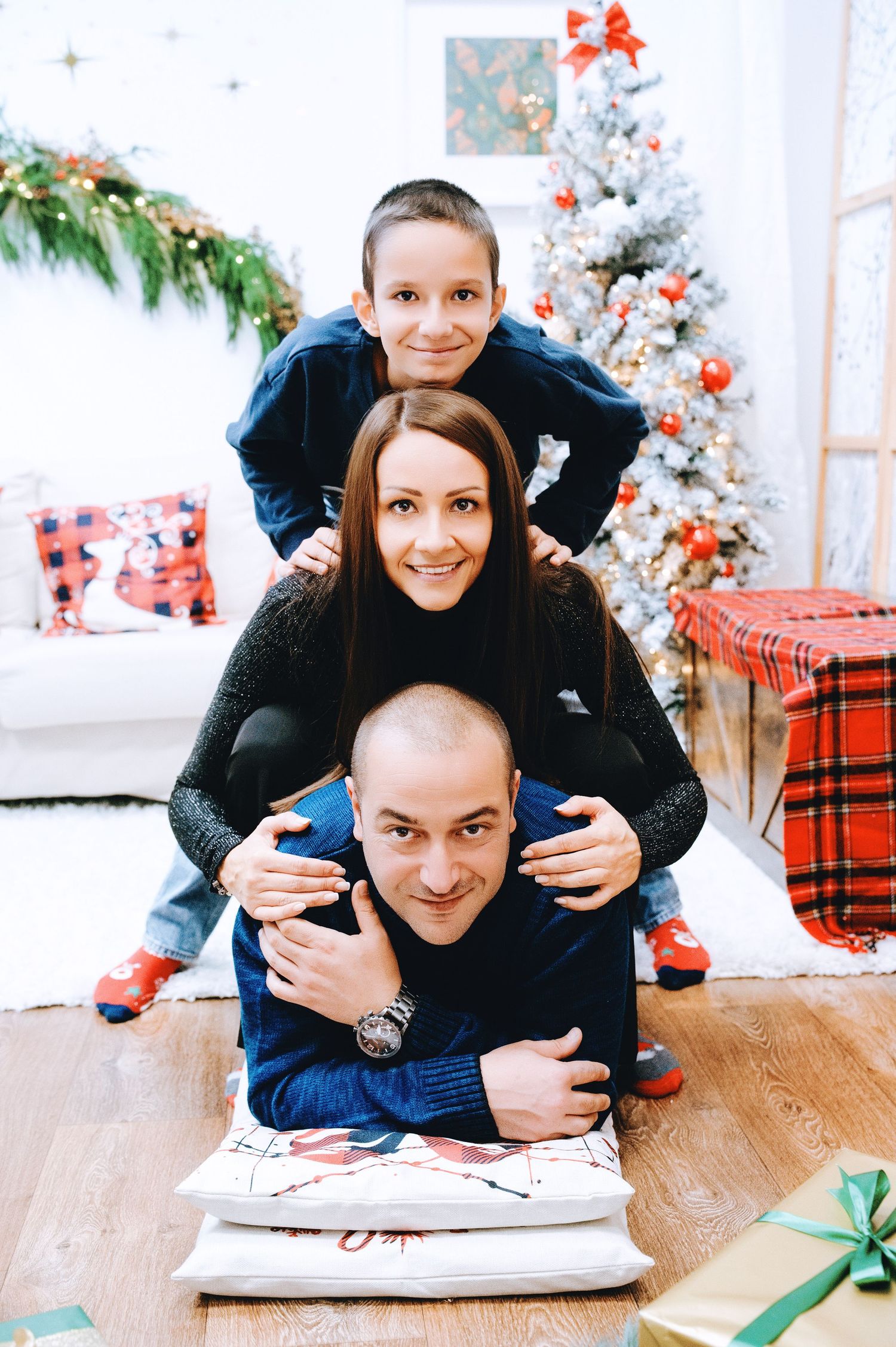 Smiling family of three stacked in a playful pyramid pose on pillows beside a decorated Christmas tree, wrapped gifts, and holiday decor.