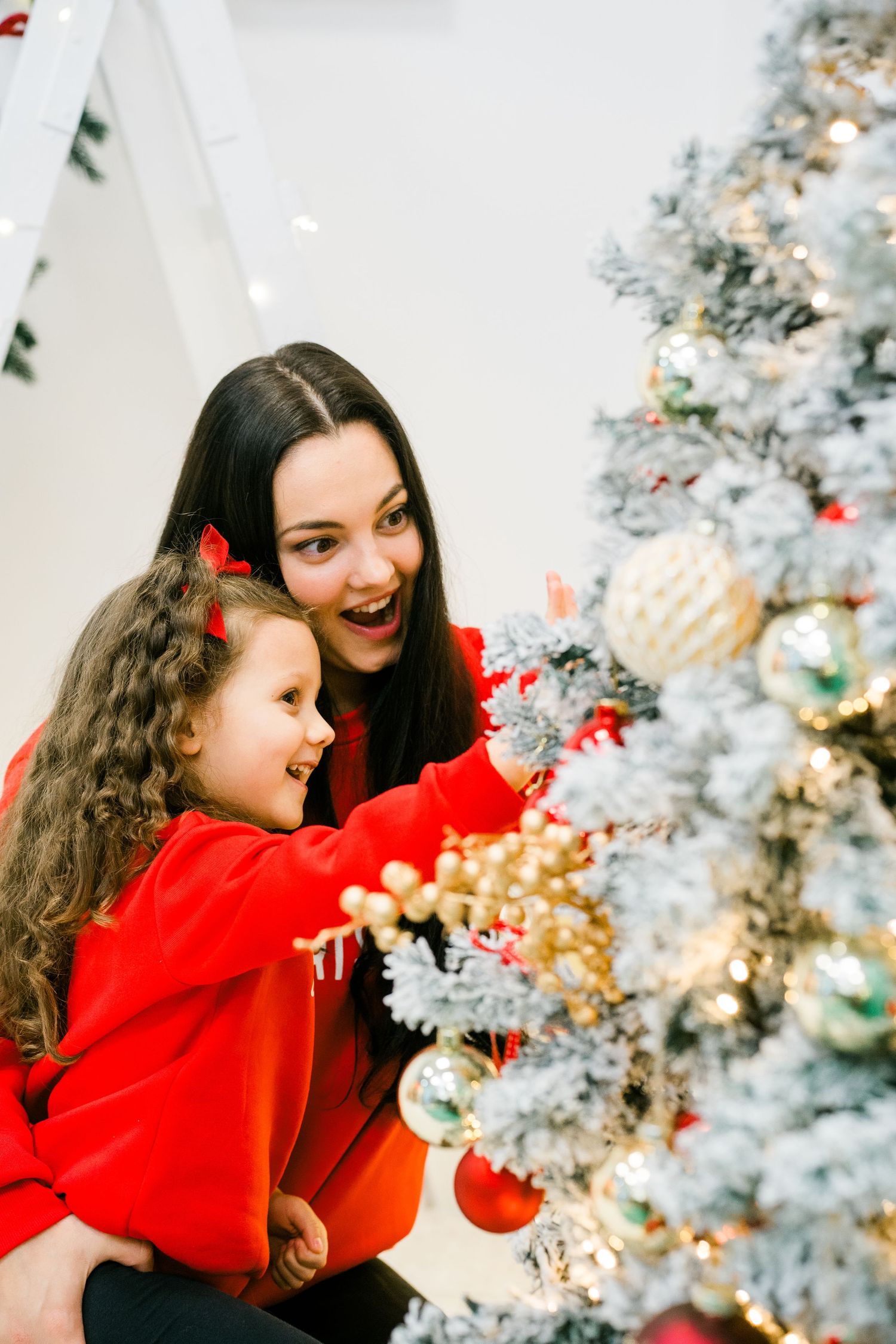 Mother and curly-haired little girl in red sweaters laughing while hanging ornaments on a frosted white Christmas tree.