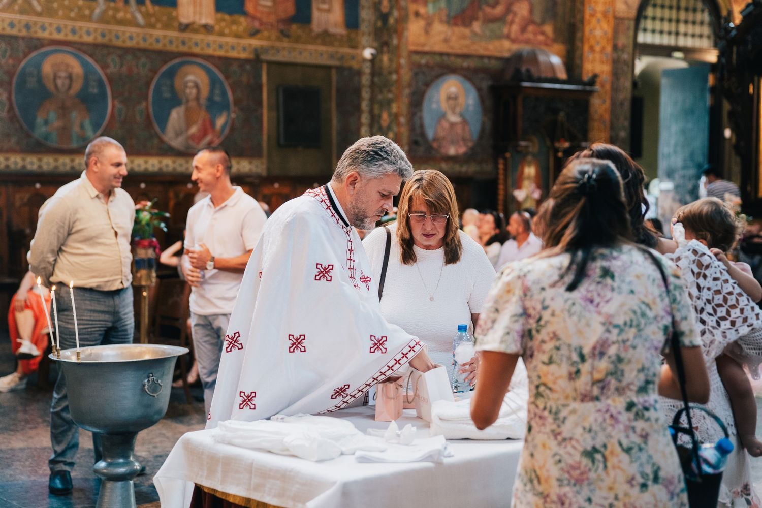 Orthodox priest in white embroidered vestments prepares a baptism table inside a church adorned with icons while guests look on.