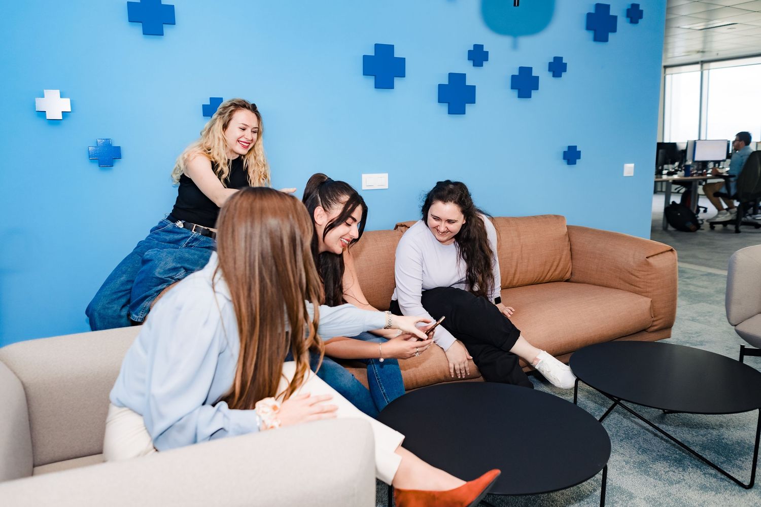 Group of women laugh while looking at a phone on cozy sofas in a vibrant blue office break area with coworkers in the background.