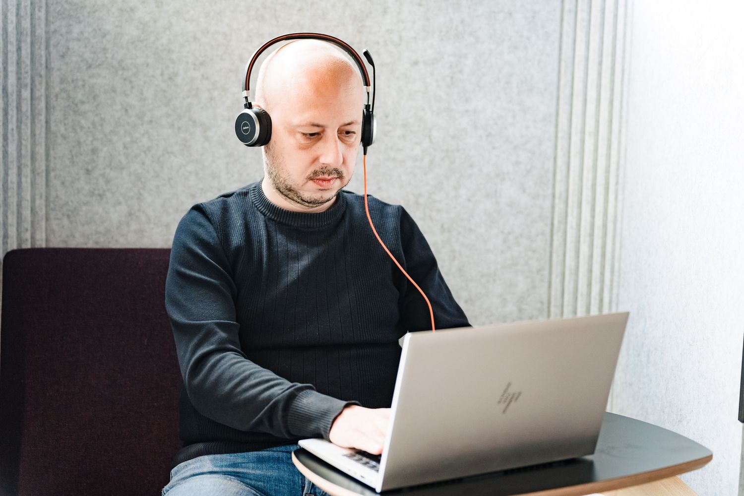 Bald man in a dark sweater and headphones types on an HP laptop at a small table in a quiet office nook.