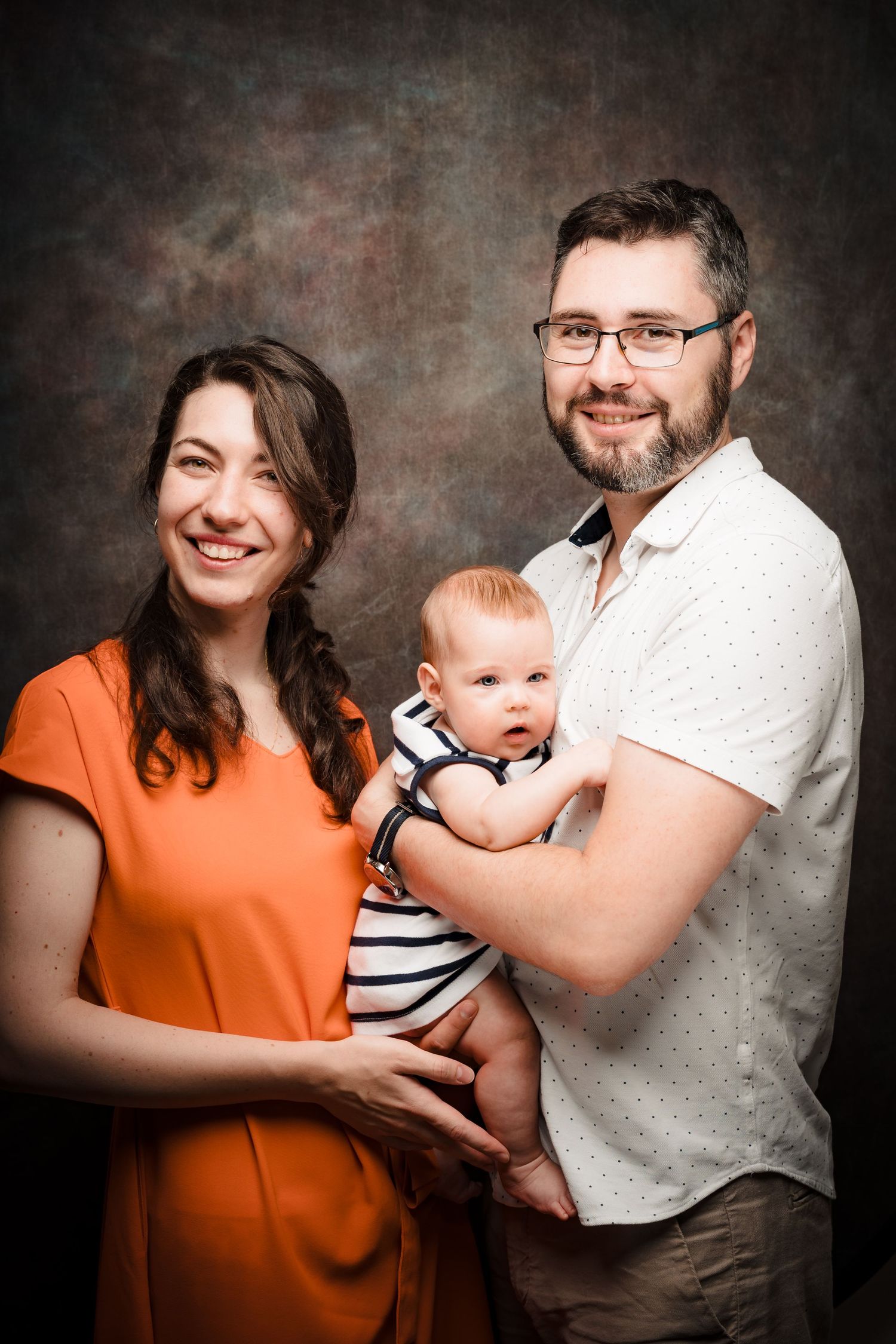 Smiling parents pose in a studio portrait, the bearded father in glasses holding their baby in a striped outfit beside the mother in orange.