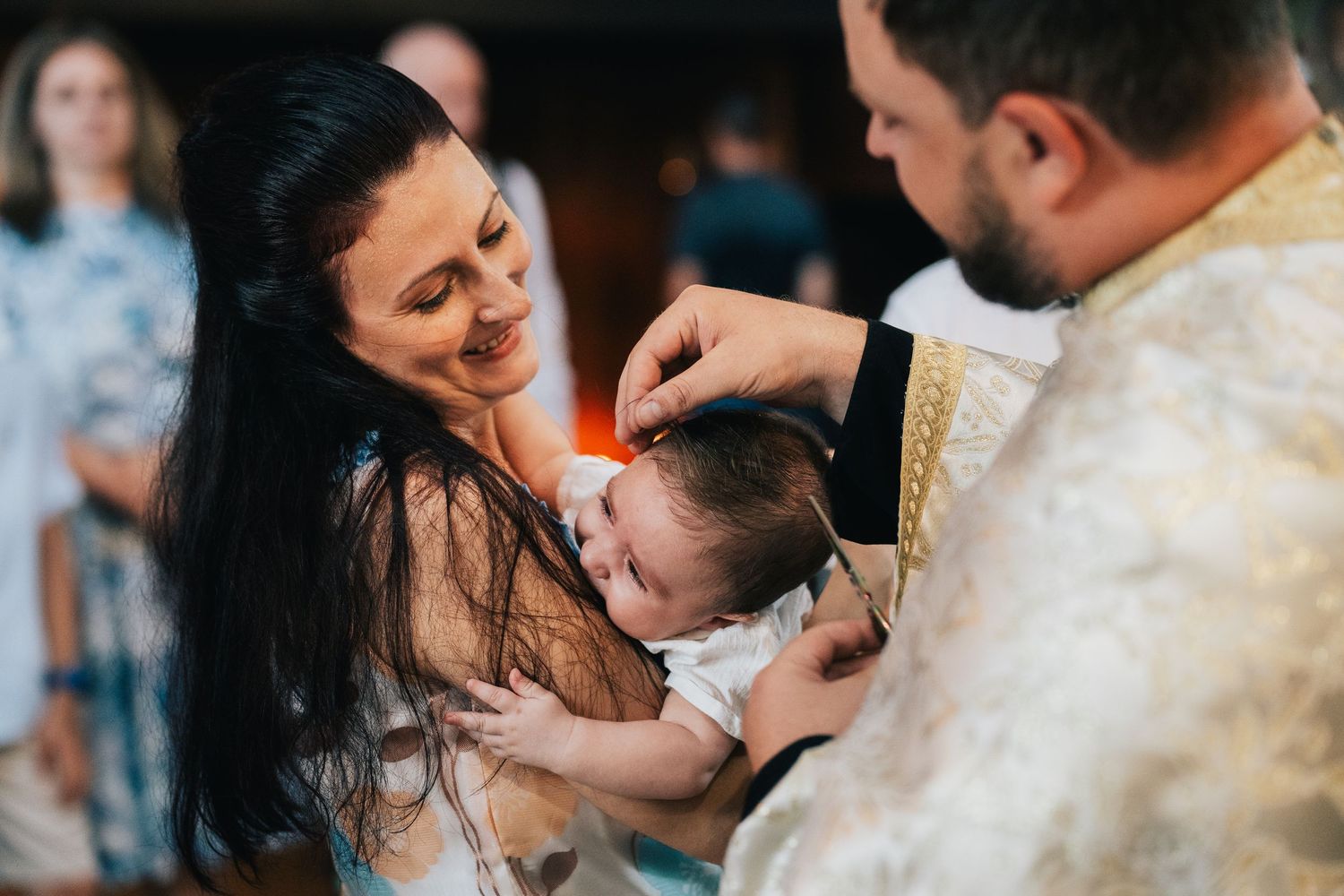 A smiling mother holds her baby as a priest in golden vestments blesses the infant's forehead during a christening ceremony.