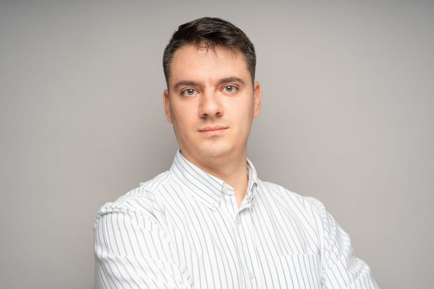 A young man with short dark hair and a striped white button-up shirt stands with crossed arms against a plain light gray background.