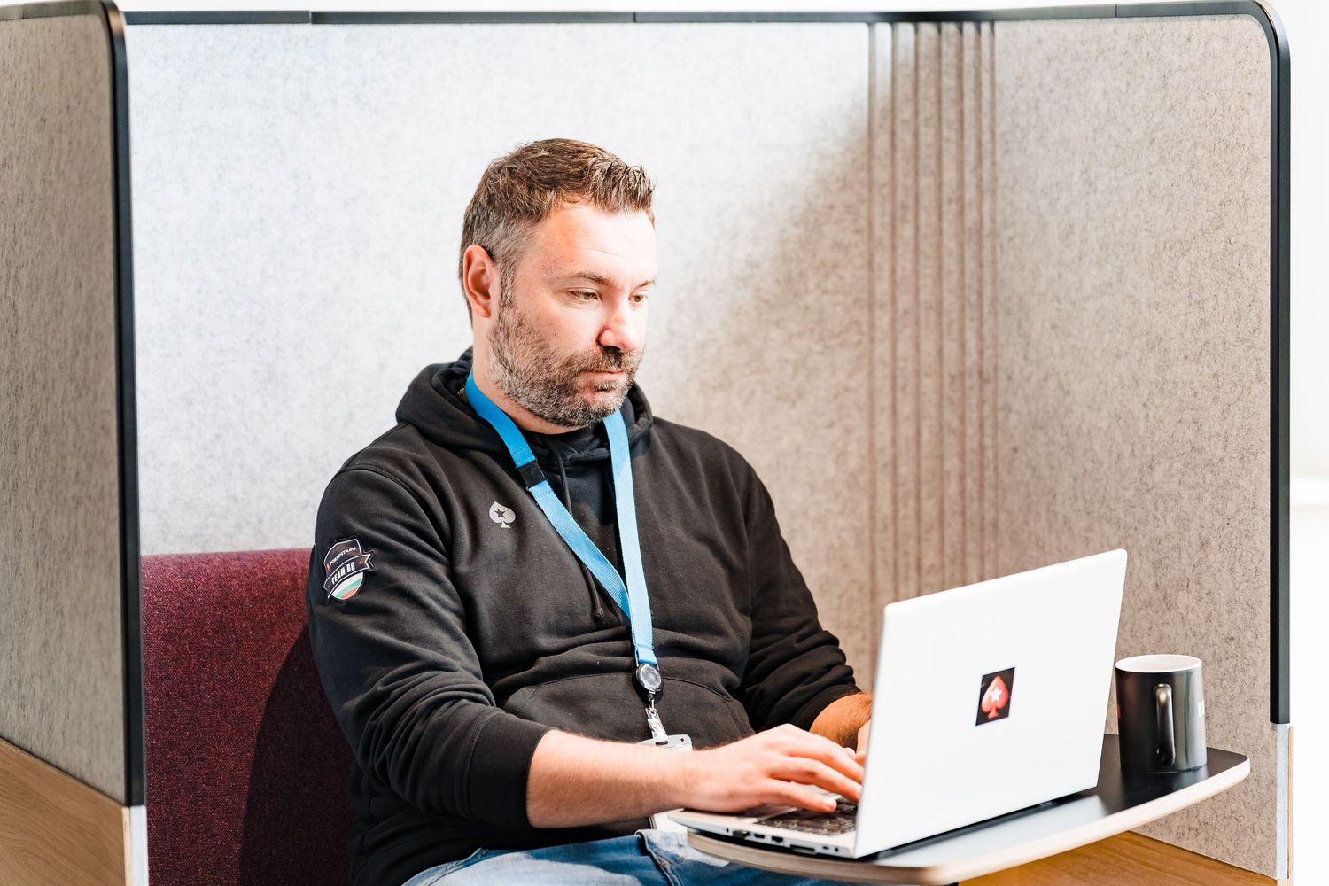 Bearded man wearing a lanyard works on a laptop in a quiet office booth with a black mug beside him.