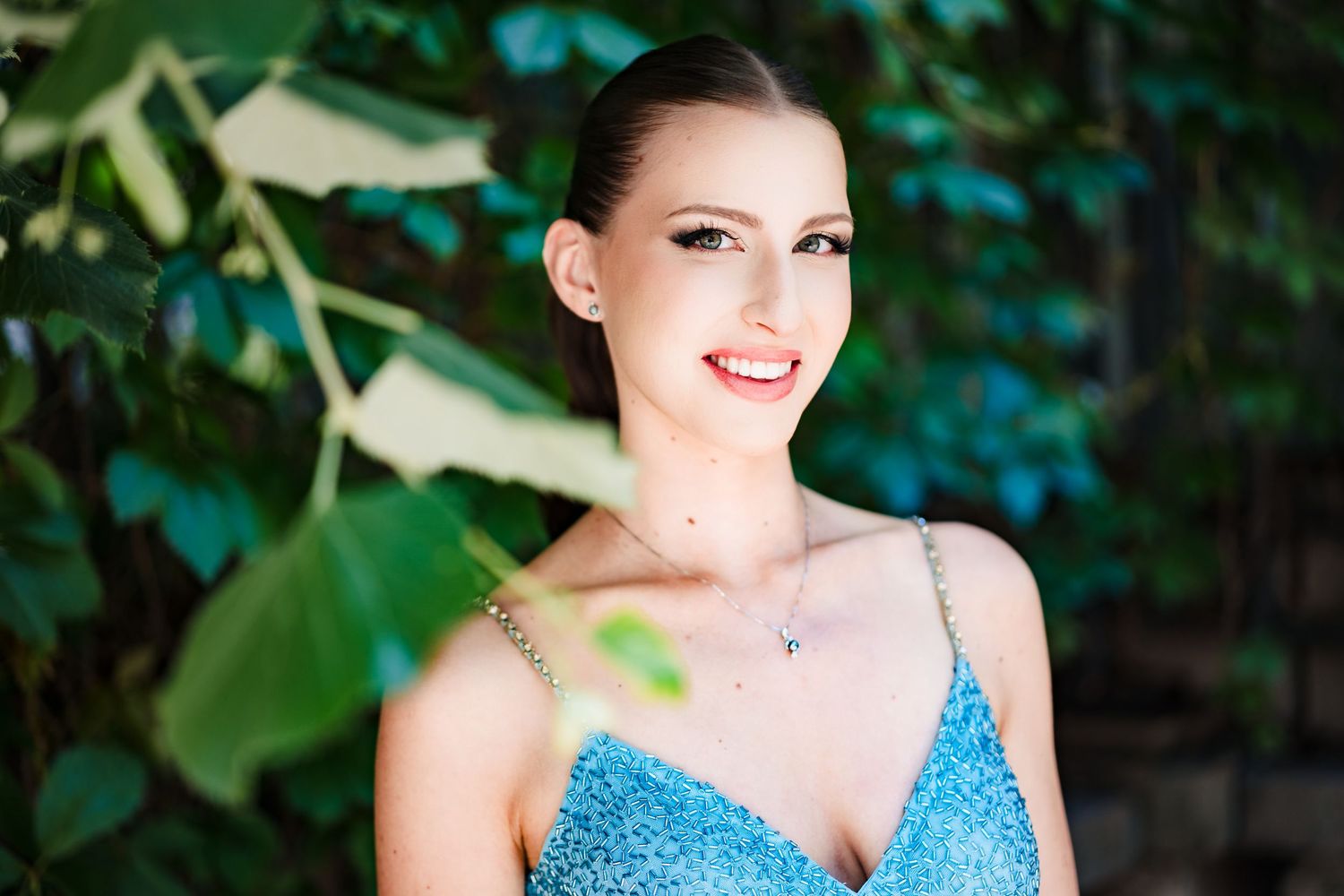 Close-up portrait of a smiling young woman in a sparkling blue dress framed by green leaves outdoors.