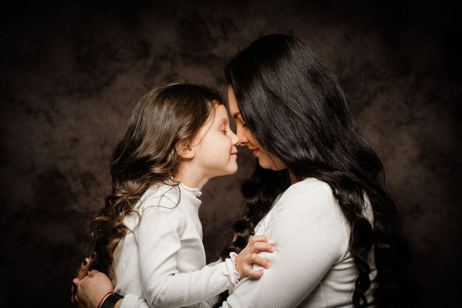 Mother and young daughter with long dark hair touch noses in a tender portrait against a dark textured studio backdrop.