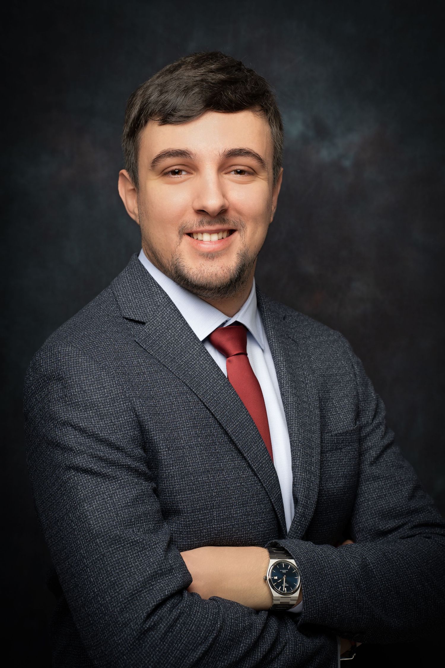 A smiling man in a gray textured suit, white shirt and red tie poses with arms crossed in a professional headshot.