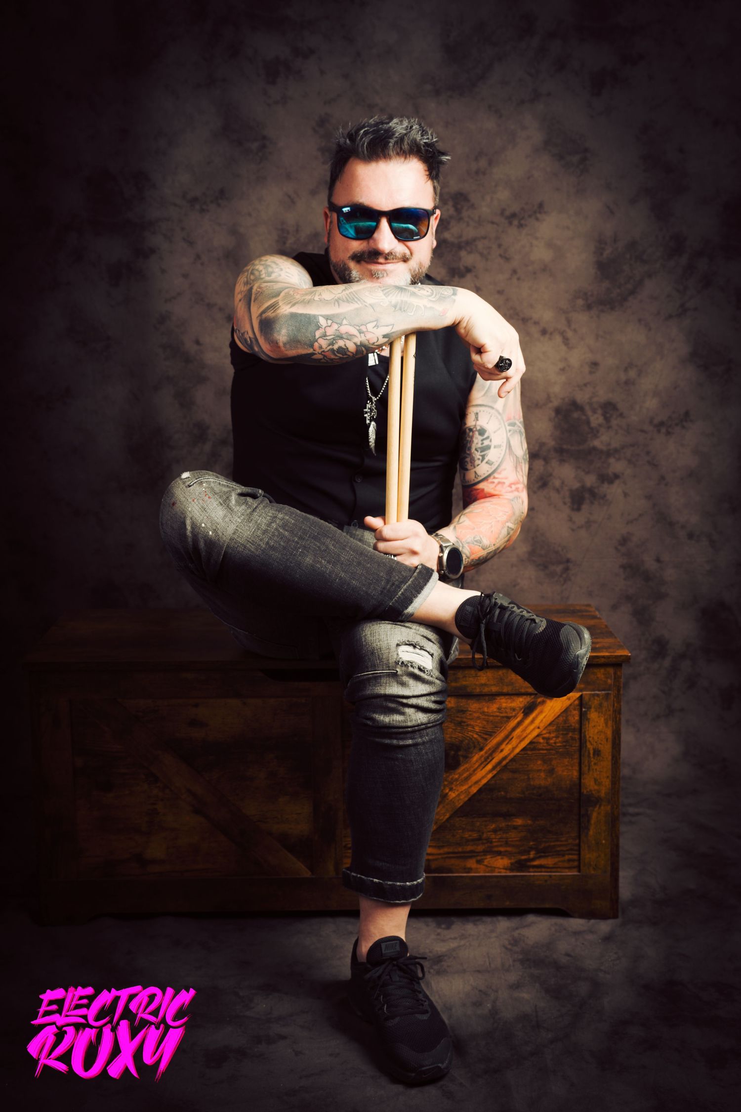 Tattooed drummer in blue mirrored sunglasses sits cross-legged on a wooden crate holding drumsticks in an Electric Roxy studio portrait.