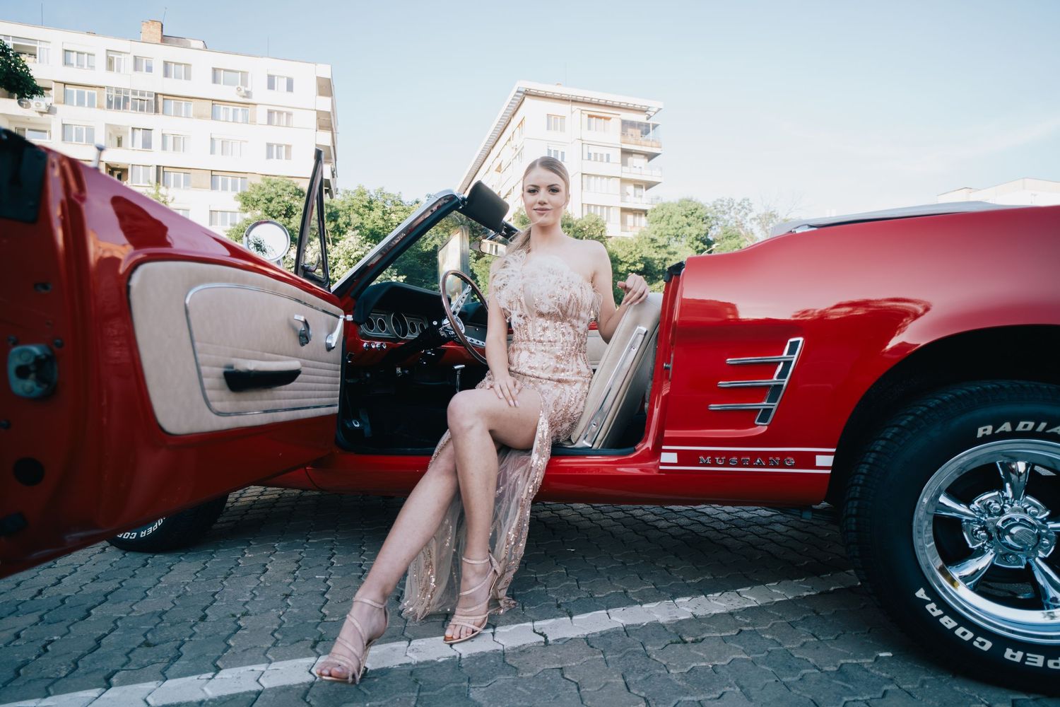 A blonde woman in a sparkly dress sits in the open door of a red vintage convertible parked on a brick-paved street.