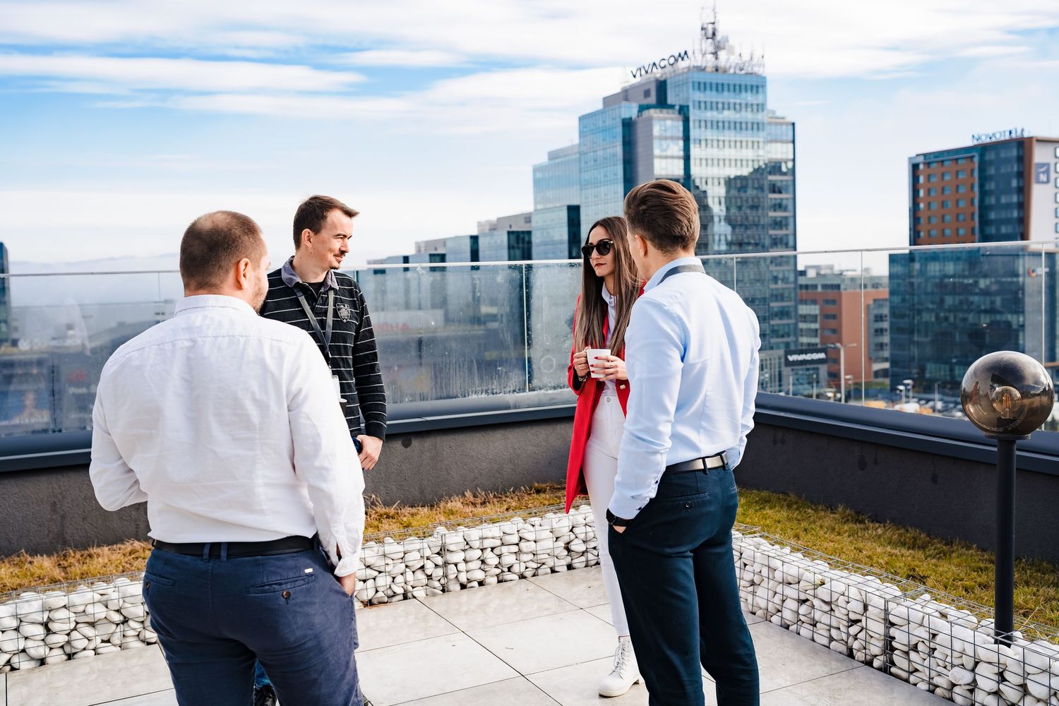 Colleagues chat on a sunny rooftop terrace with a city skyline and modern office buildings in the background.