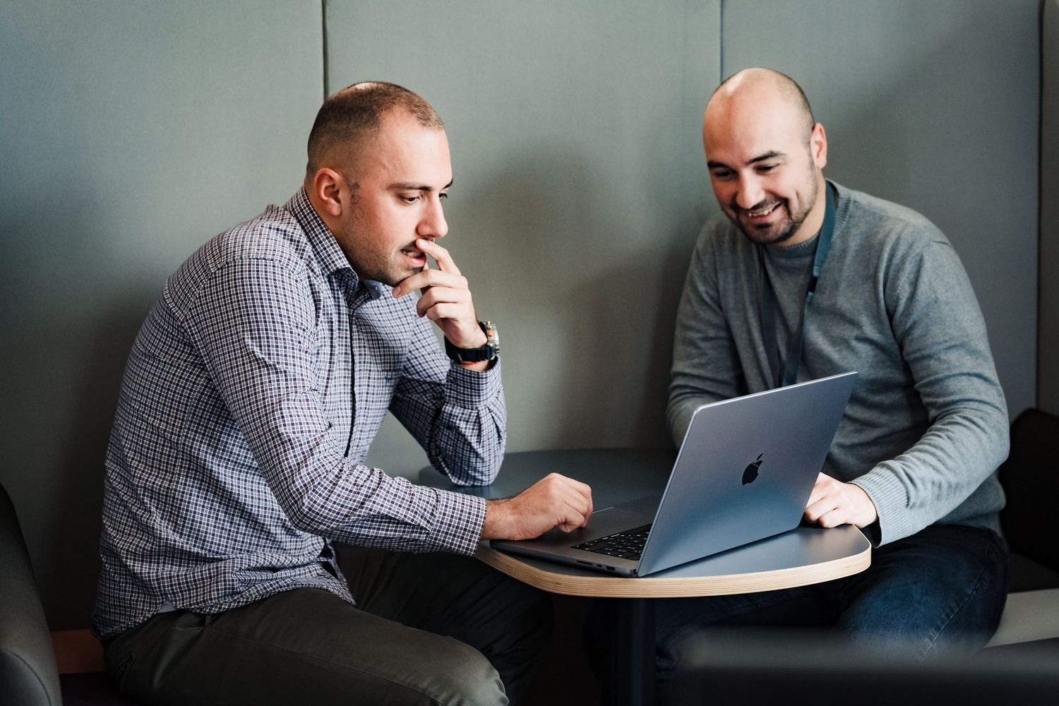 Two smiling colleagues sit close together and review work on a MacBook laptop in a casual office lounge.