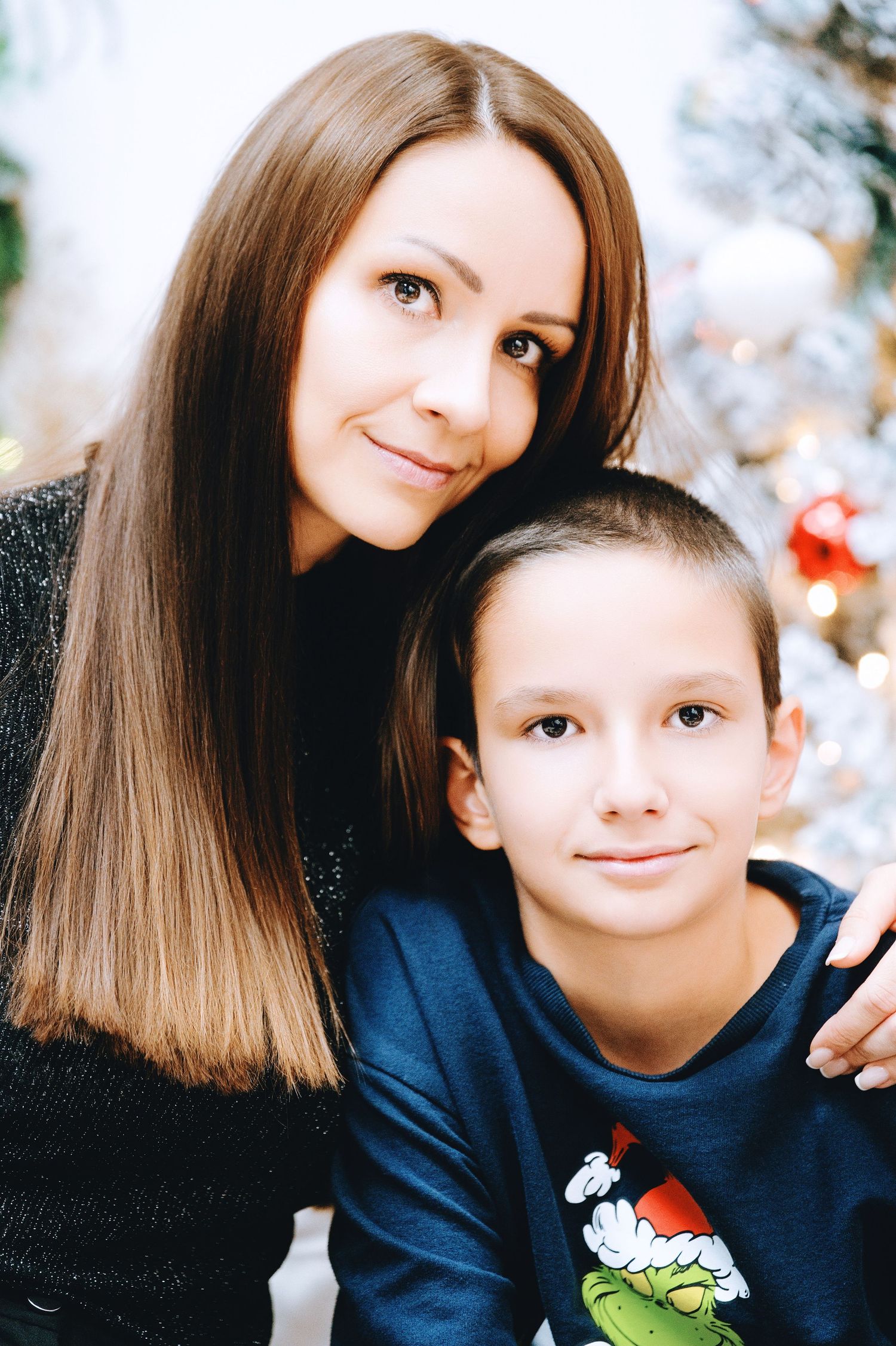 A smiling woman with long brown hair hugs a boy in a Grinch Christmas sweater beside a decorated tree with ornaments and lights.