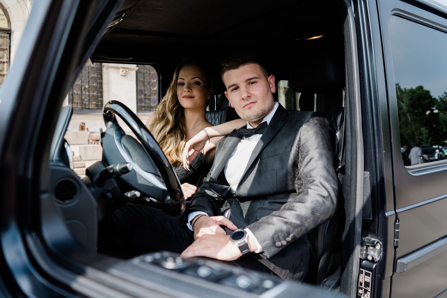 A young man in a gray suit and bow tie sits in a black SUV beside a blonde woman in an elegant evening look.