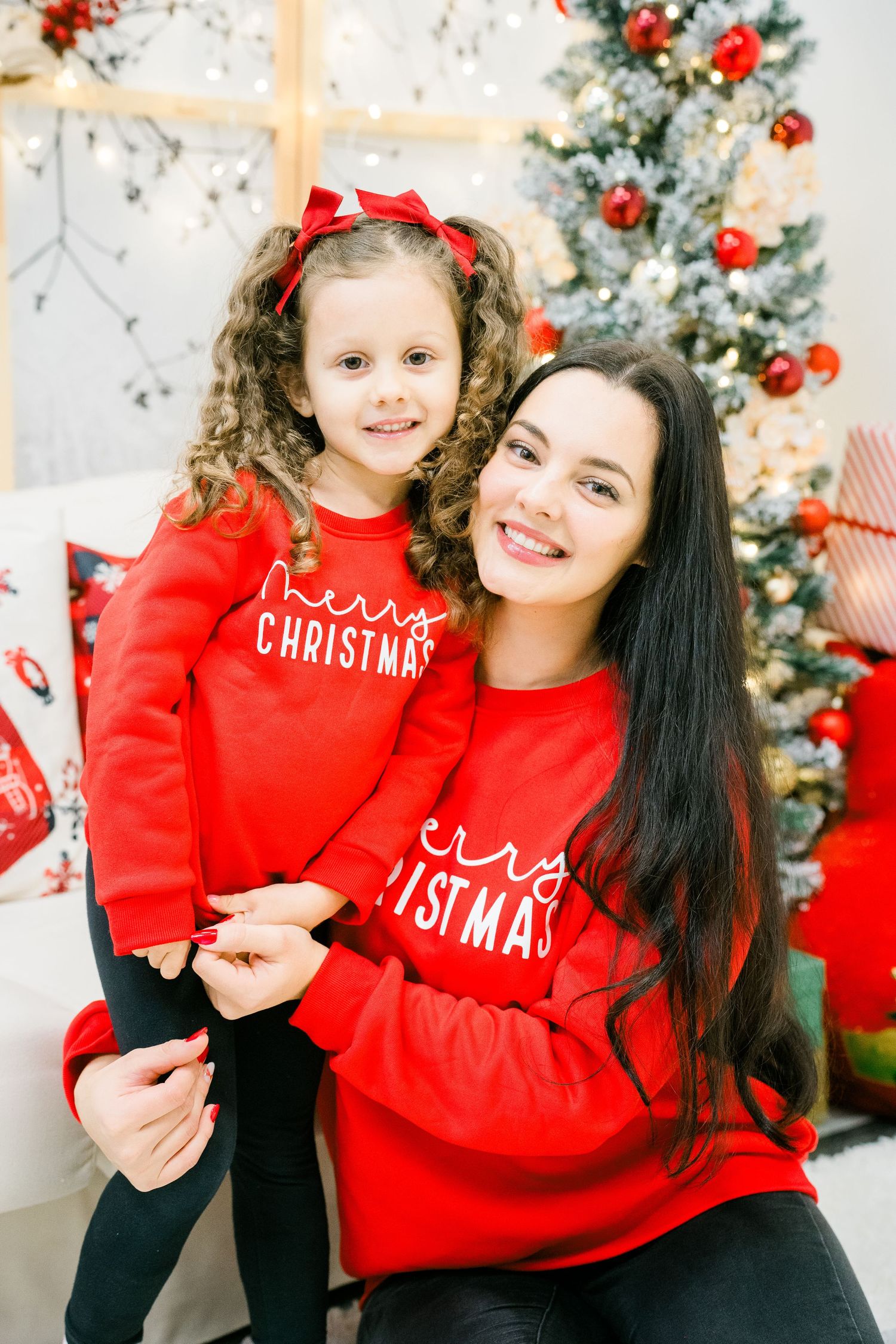 Smiling mother and young daughter in matching red Christmas sweatshirts hugging in front of a decorated Christmas tree.