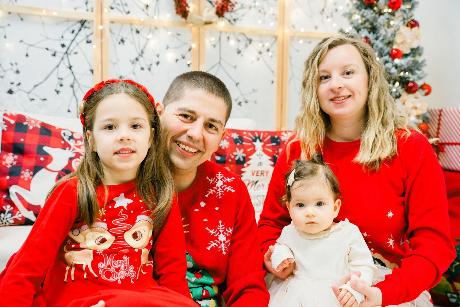 Parents and two daughters in red holiday outfits smile together near a Christmas tree in a bright festive family portrait.