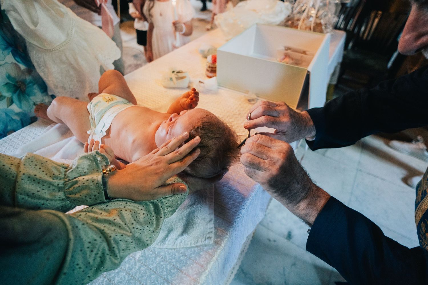 A baby lies on a white cloth during an Orthodox baptism as a priest's hands anoint the child with holy oil.