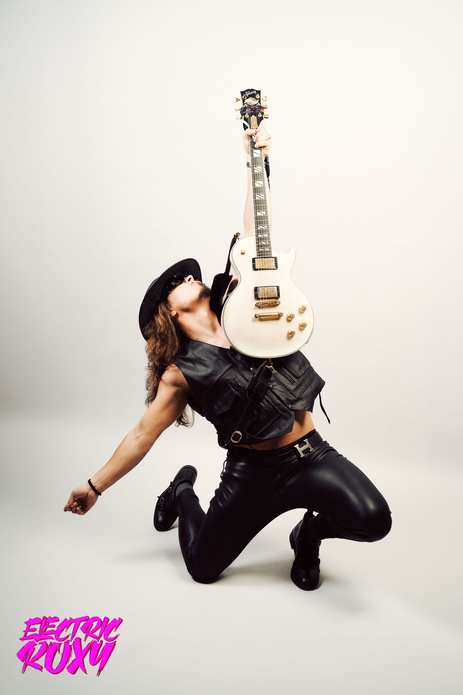 Rock musician in leather vest and pants kneels while lifting a white electric guitar high above his face against a plain studio backdrop.