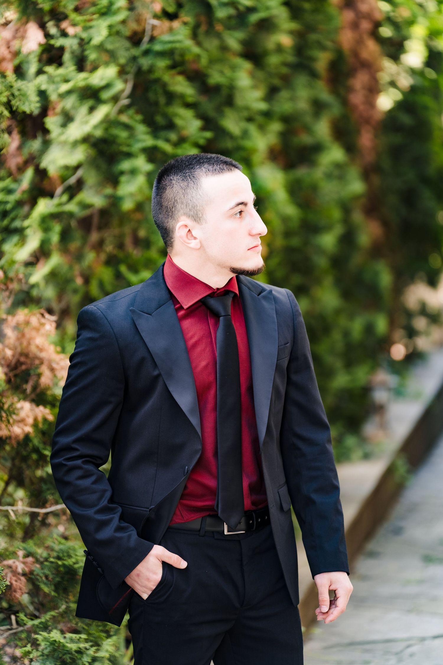Young man in a black blazer, burgundy shirt and tie stands with a hand in his pocket beside lush green foliage.