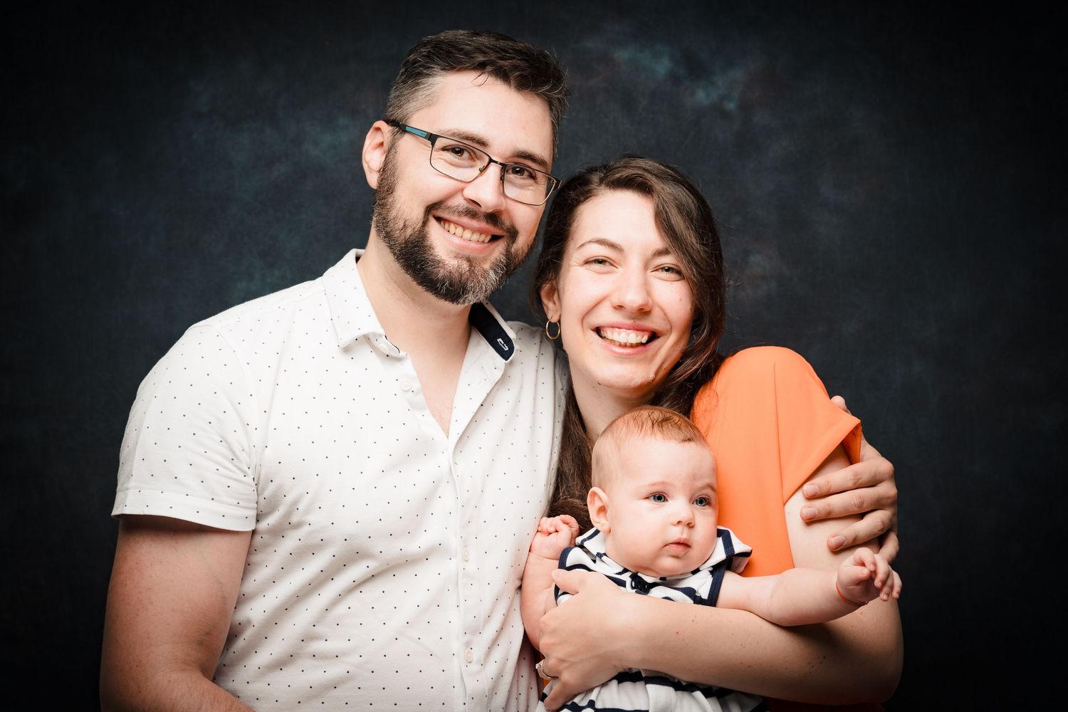 A smiling couple holds their baby between them in a close studio family portrait against a dark blue background.