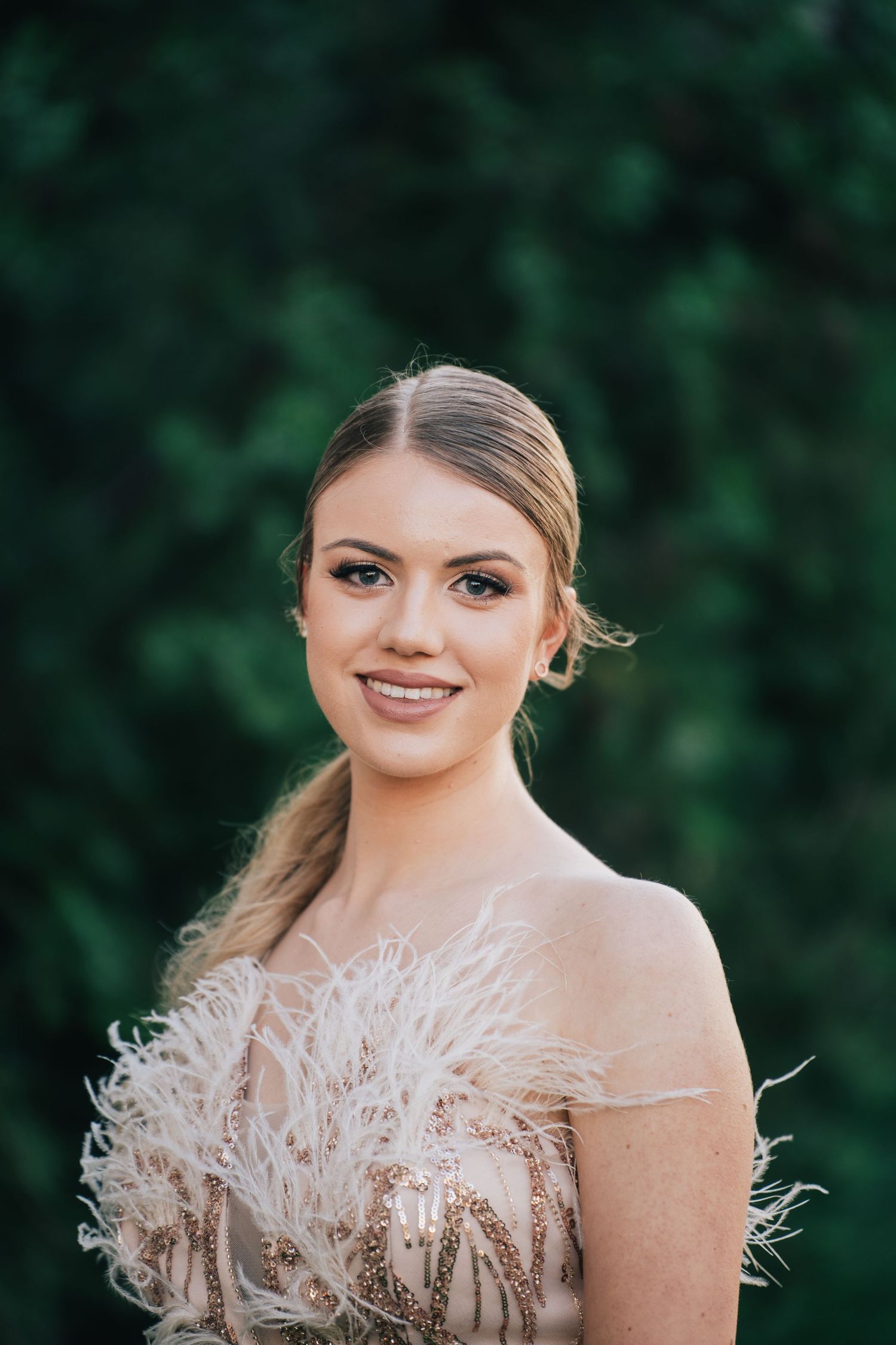 Portrait of a smiling young woman with a sleek ponytail wearing a feathered, sequined blush dress against green foliage.