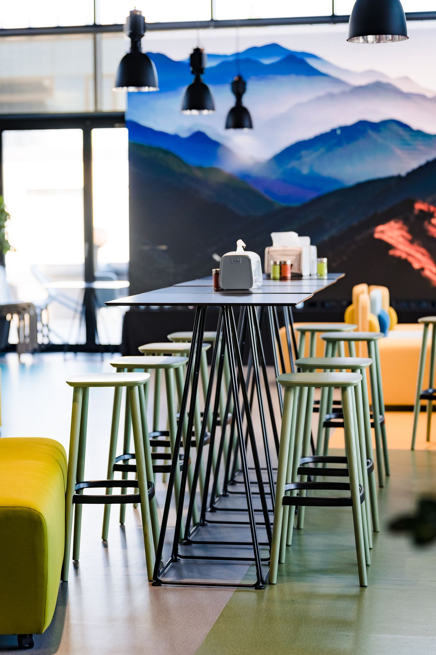 Tall bar table with green stools in a bright modern cafe with pendant lights and a mountain landscape mural.