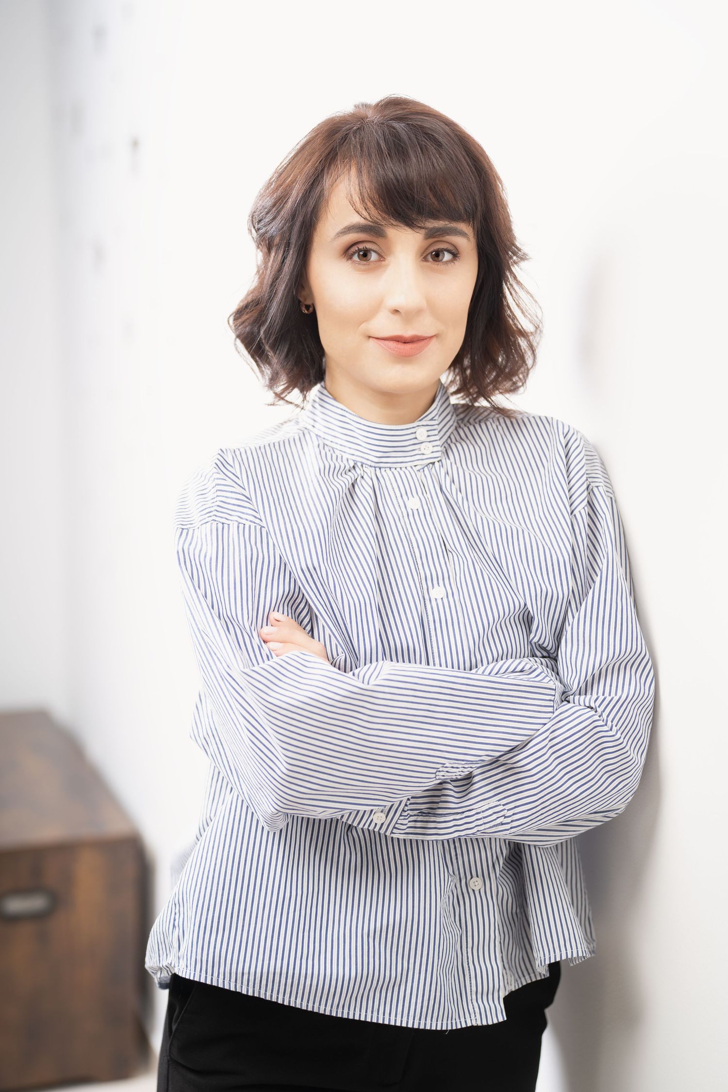 Woman with short wavy brown hair and a striped blouse stands with crossed arms against a white wall in a studio portrait.