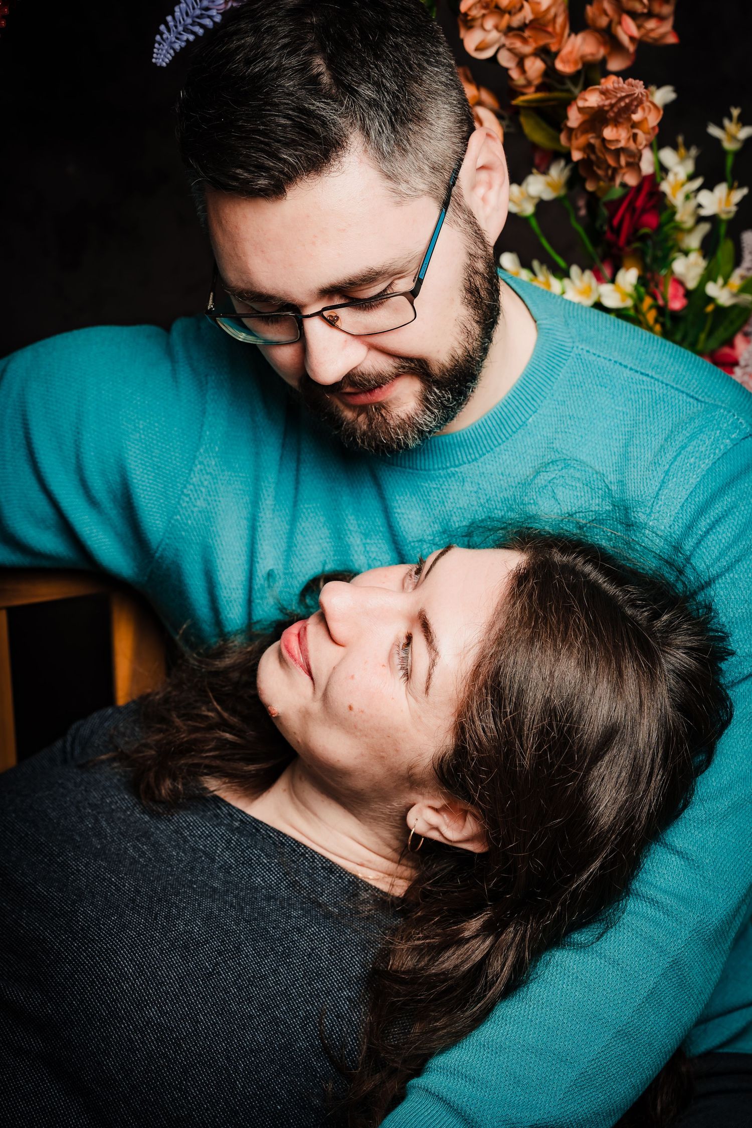 A bearded man in glasses and teal sweater gazes down at a woman resting on his lap, with autumn flowers behind them in a couple portrait.