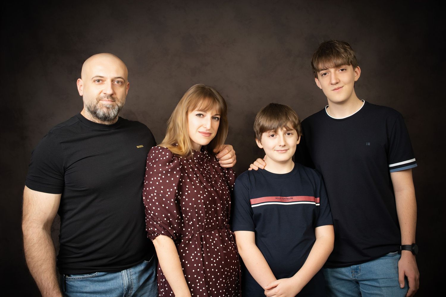 A family portrait of a bald bearded man, a woman and two boys in dark clothing posing together against a brown studio backdrop.