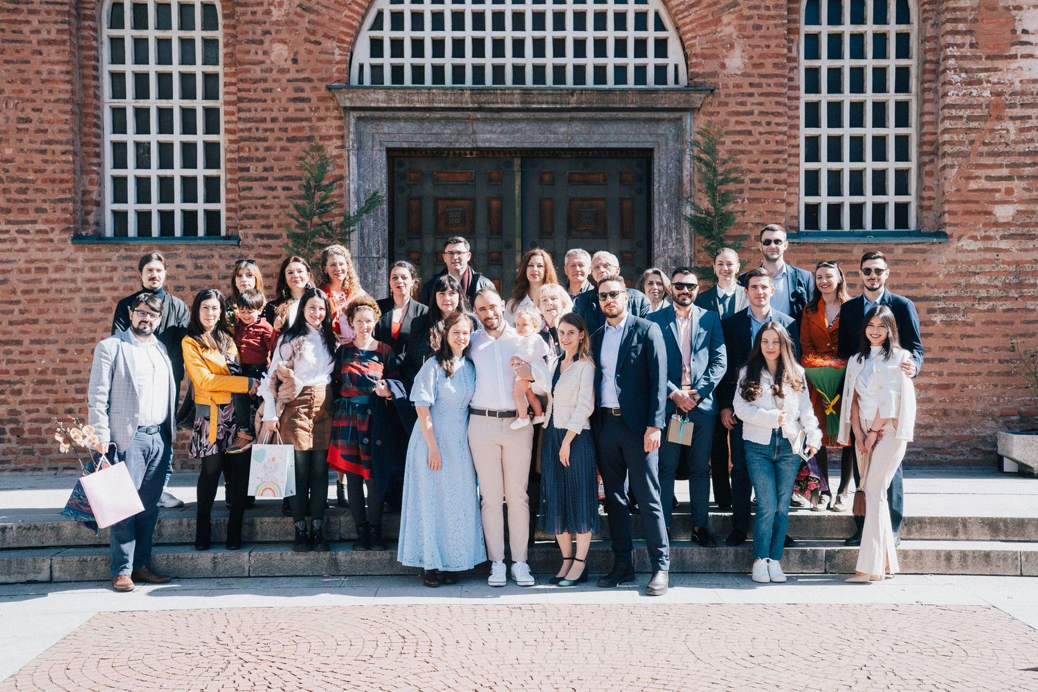 A large group of family and friends in elegant outfits poses together on the steps of a red brick church after a baptism.