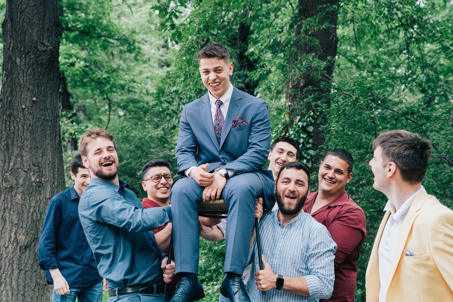 A group of cheerful men lift a smiling groom in a blue suit into the air while celebrating outdoors among green trees.