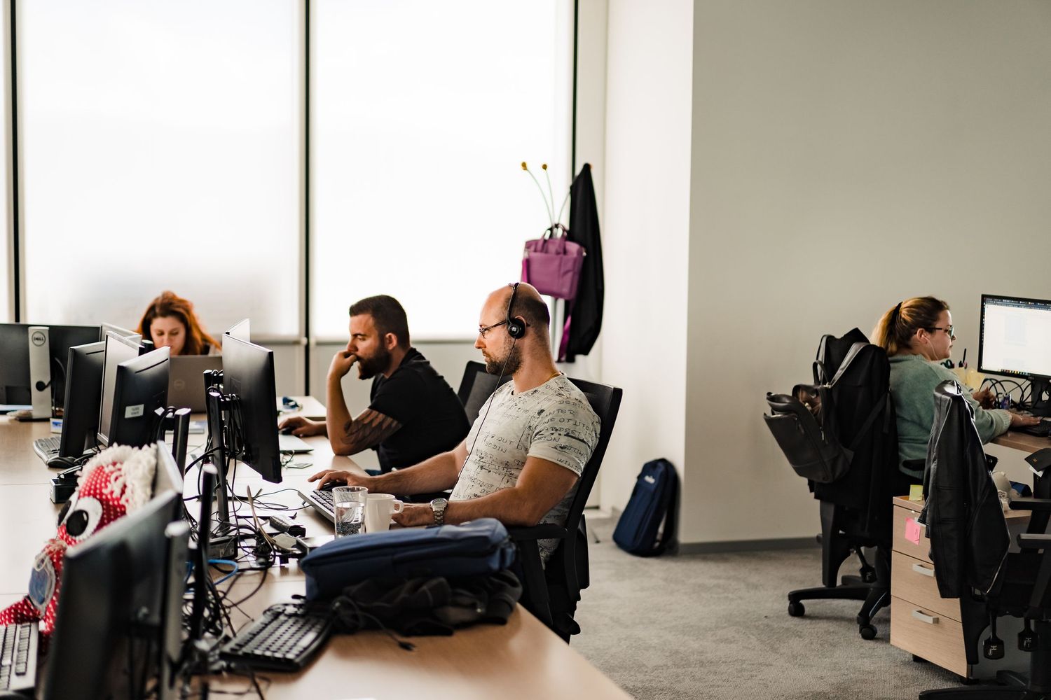 Employees wearing headsets and working at computer monitors at long desks in a bright open-plan office with large windows.