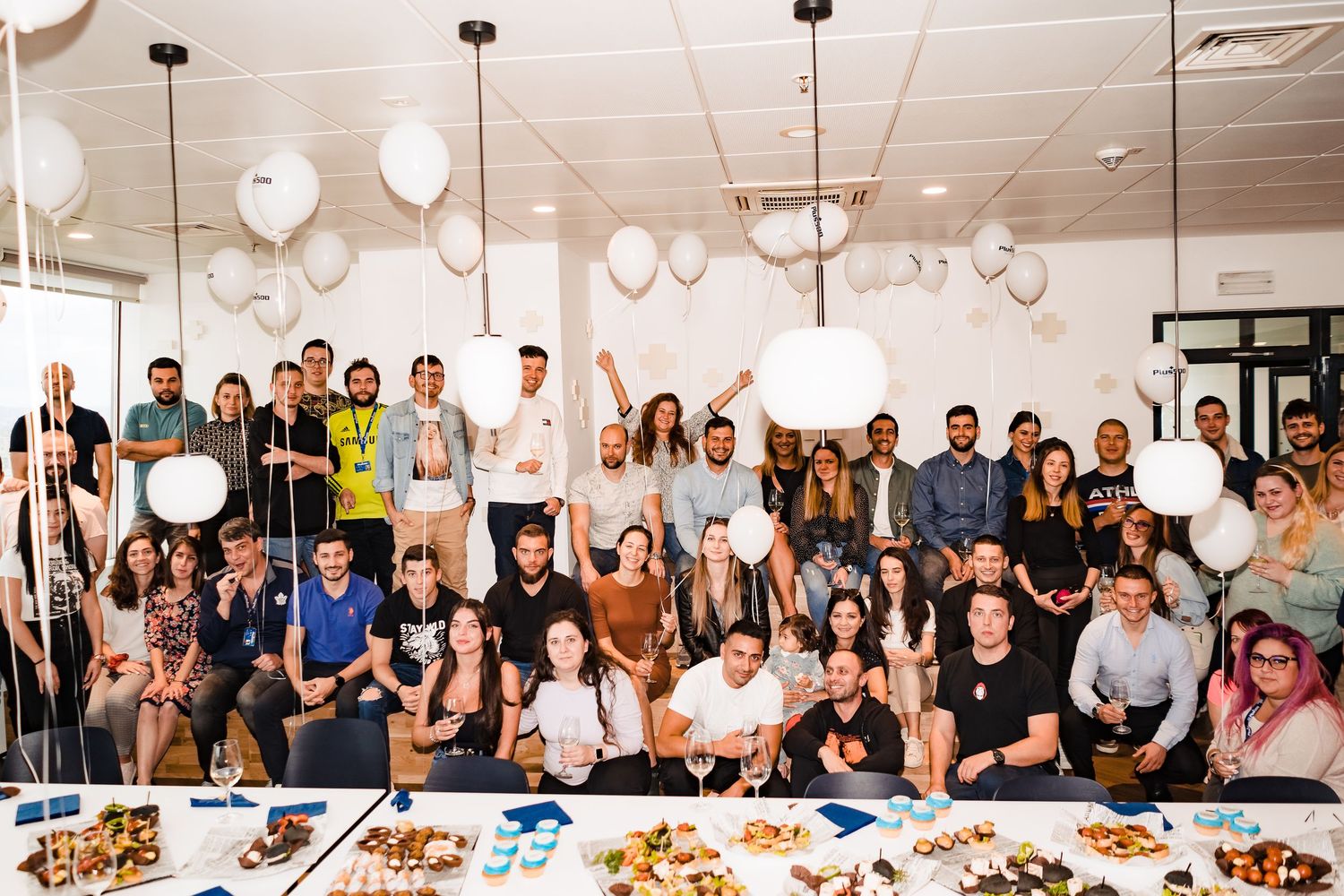 Large group photo of smiling coworkers posing together at a party with white balloons, pendant lights and tables of snacks.