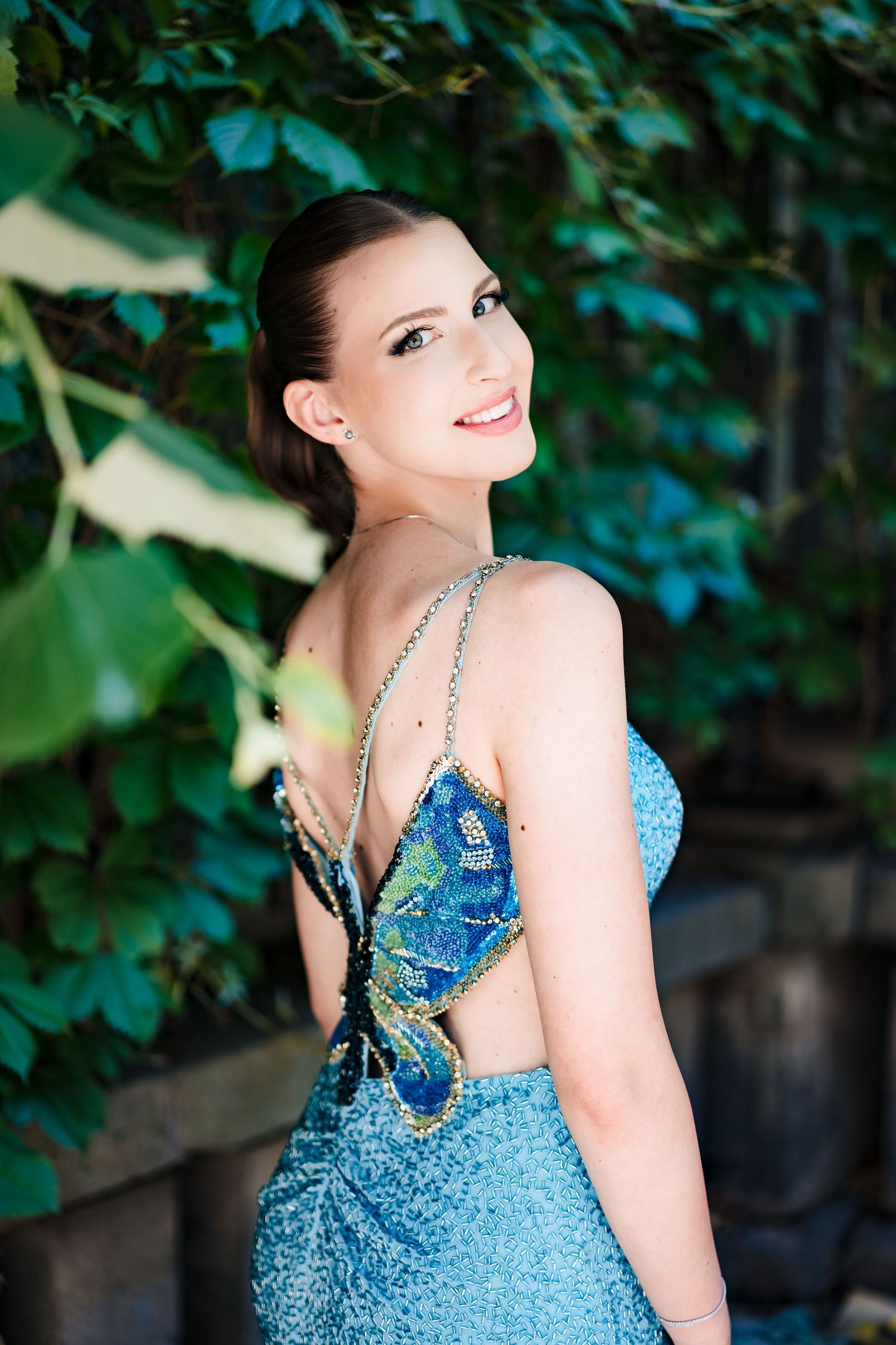 Smiling young woman looks over her shoulder, showing the butterfly-style open back of her sparkling blue dress among green foliage.