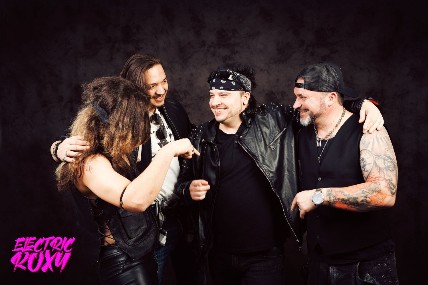 Four Electric Roxy band members laugh and embrace in a playful group pose against a dark studio background, wearing leather and bandanas.
