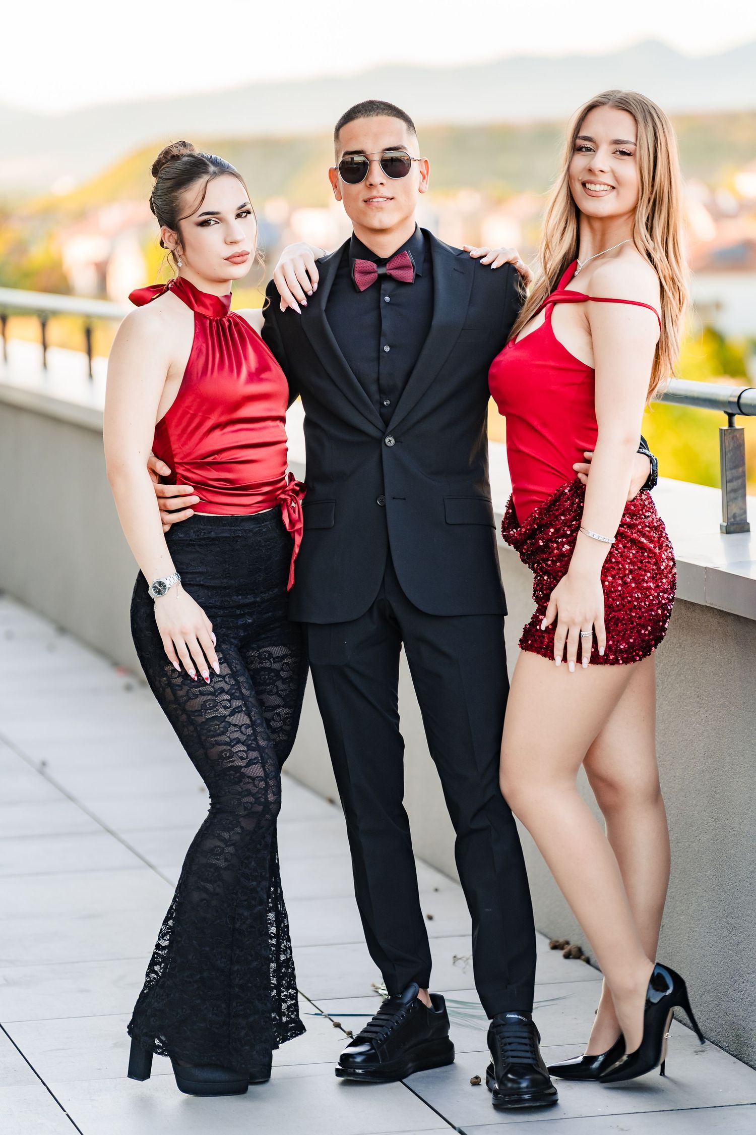 Man in a black suit, bow tie and sunglasses stands between two smiling women in red outfits on a sunny terrace.