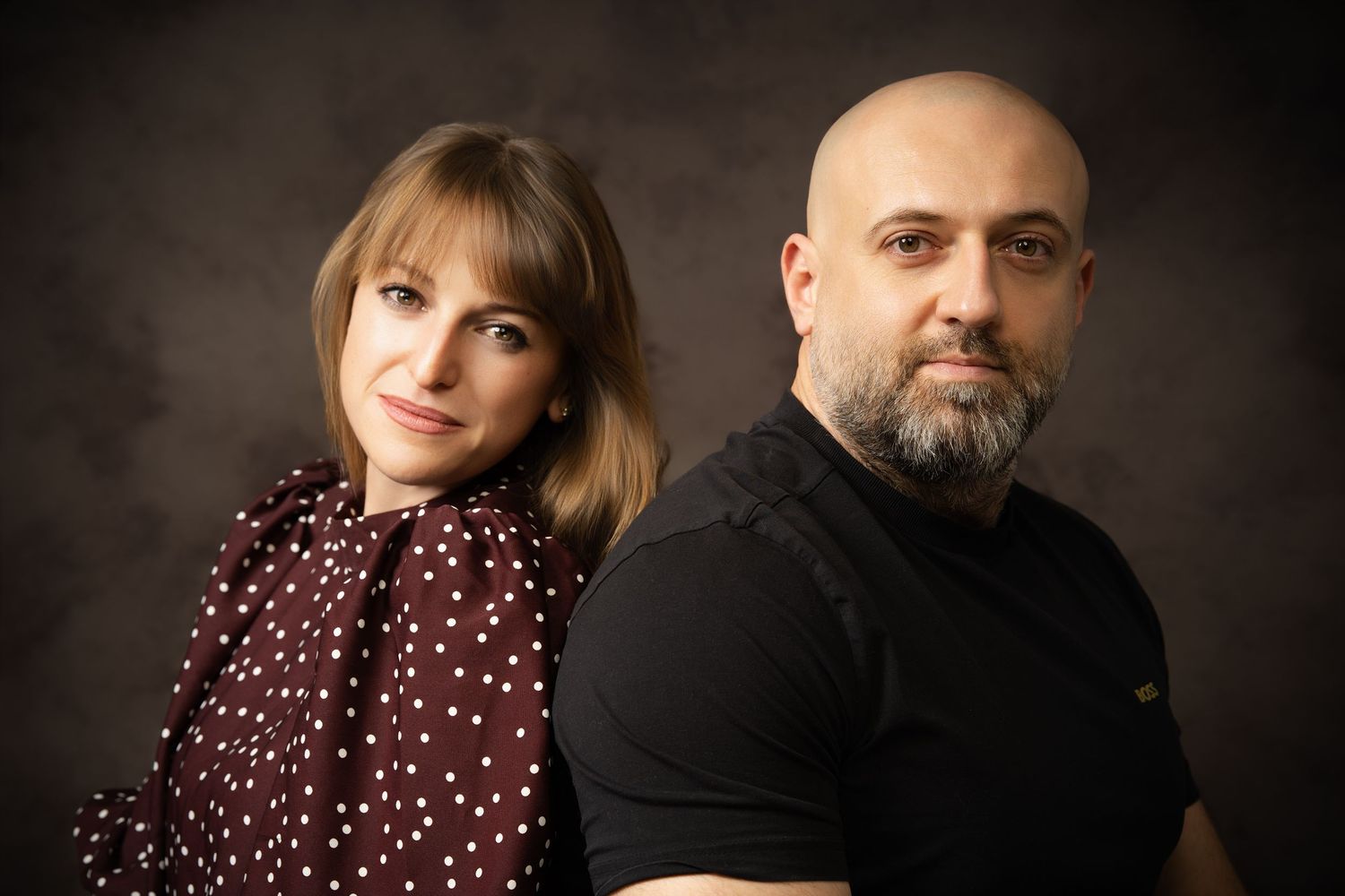 A woman in a burgundy polka dot blouse and a bald bearded man in black pose side by side in a moody studio portrait.