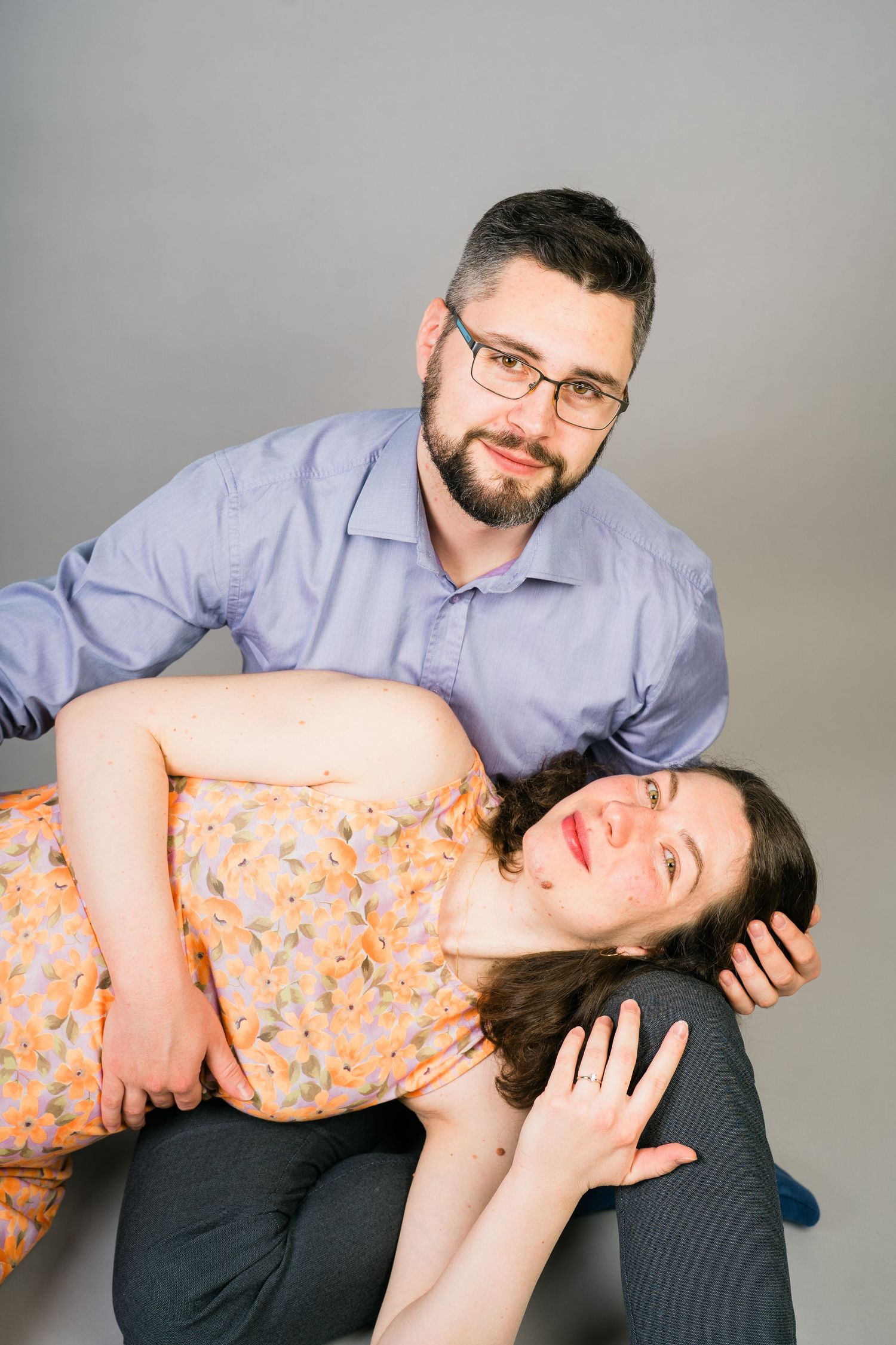 Pregnant woman in a floral dress lies across her bearded partner's lap as he smiles in a relaxed maternity photo session.