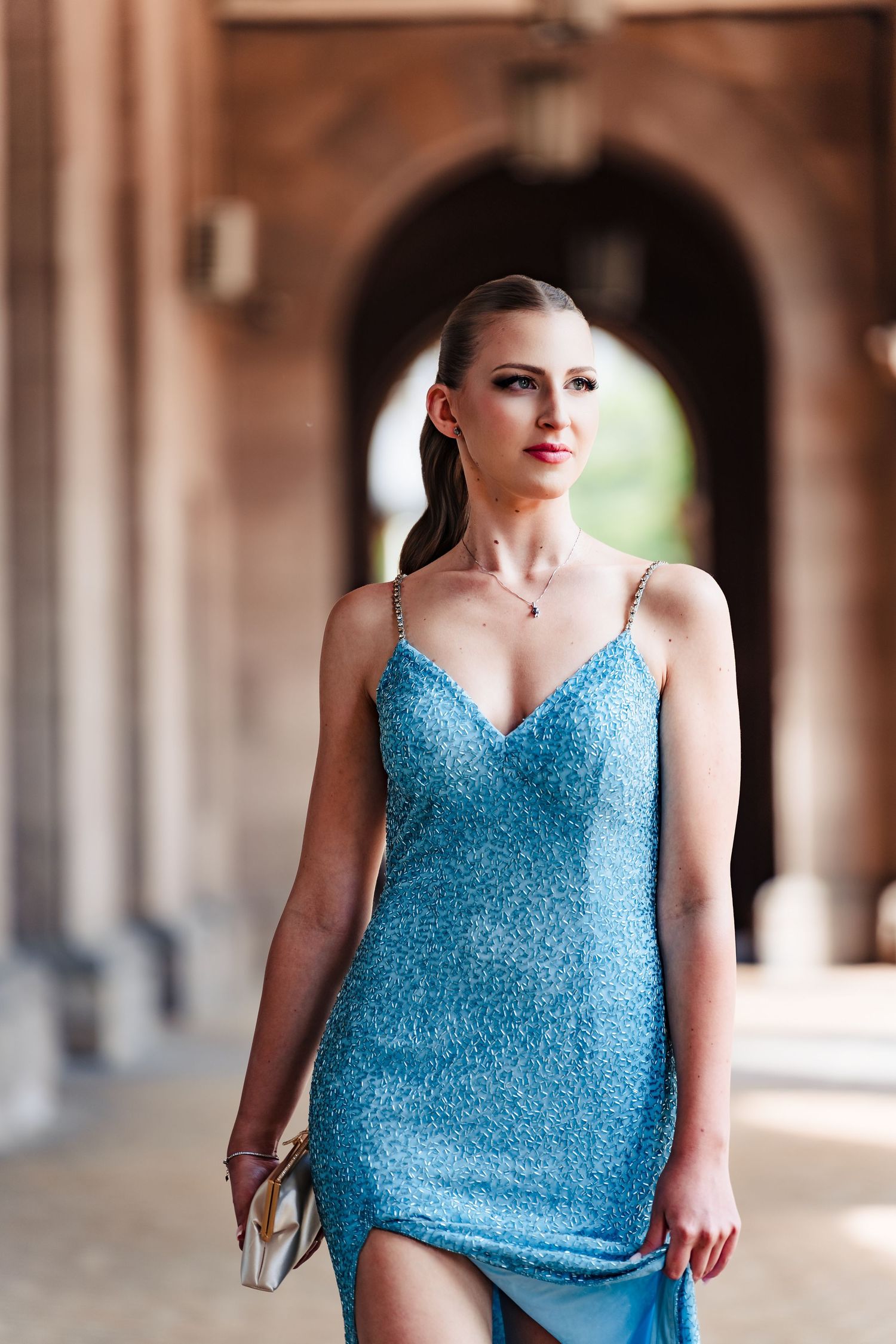 A woman with slicked-back hair walks beneath a stone archway in a sparkling blue sequin dress, holding a small clutch.