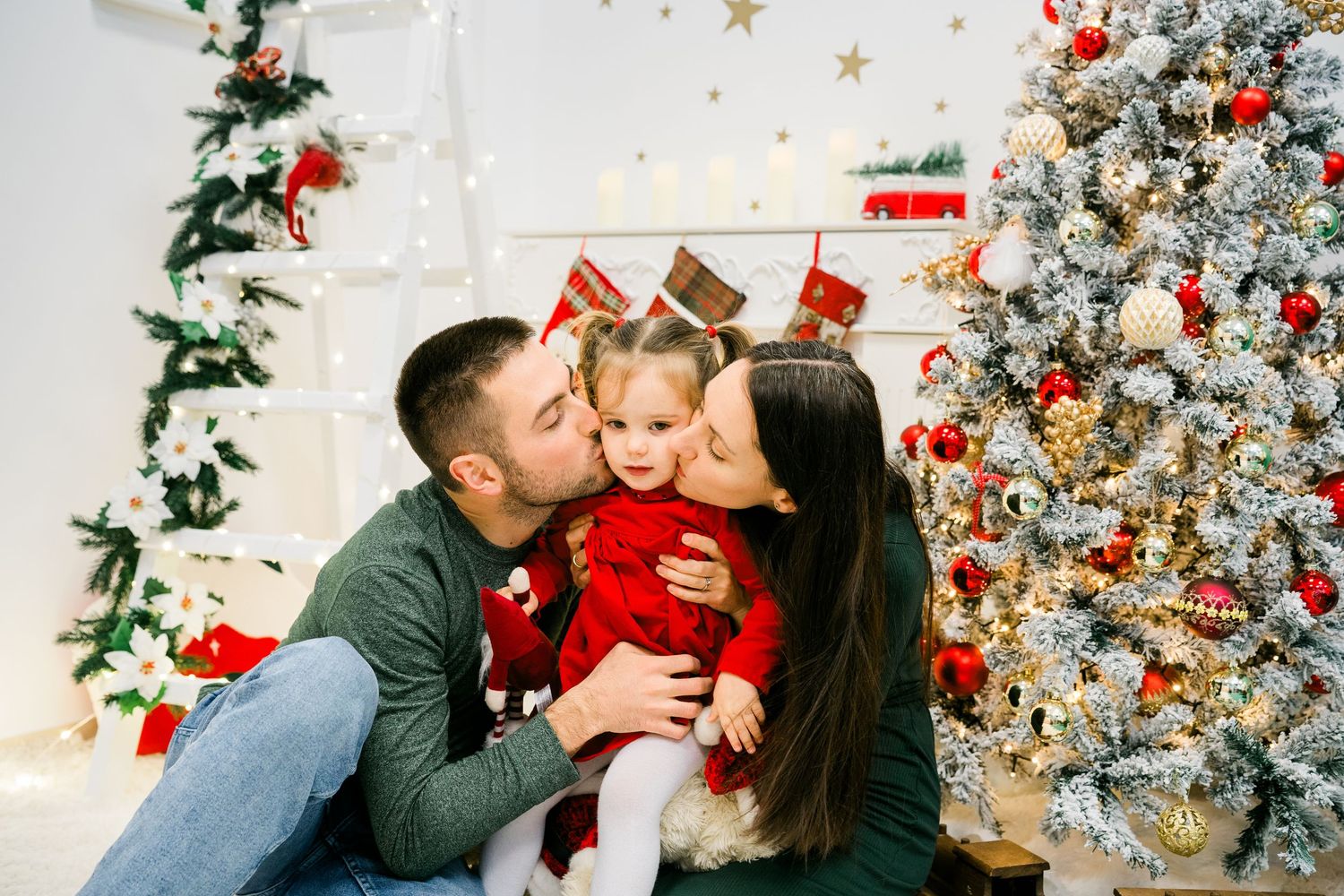 Mother and father kiss their toddler daughter in a red dress on both cheeks beside a decorated Christmas tree.