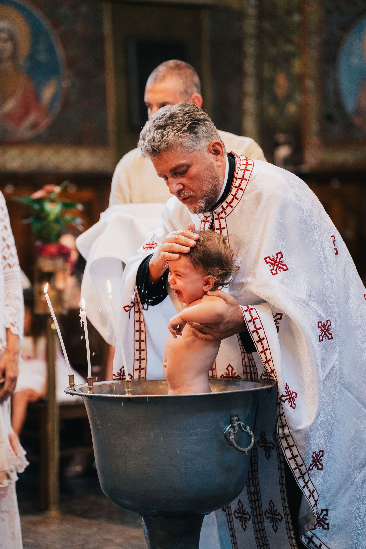 An Orthodox priest in embroidered white robes gently pours water over a baby's head in a baptismal font during a christening ceremony.