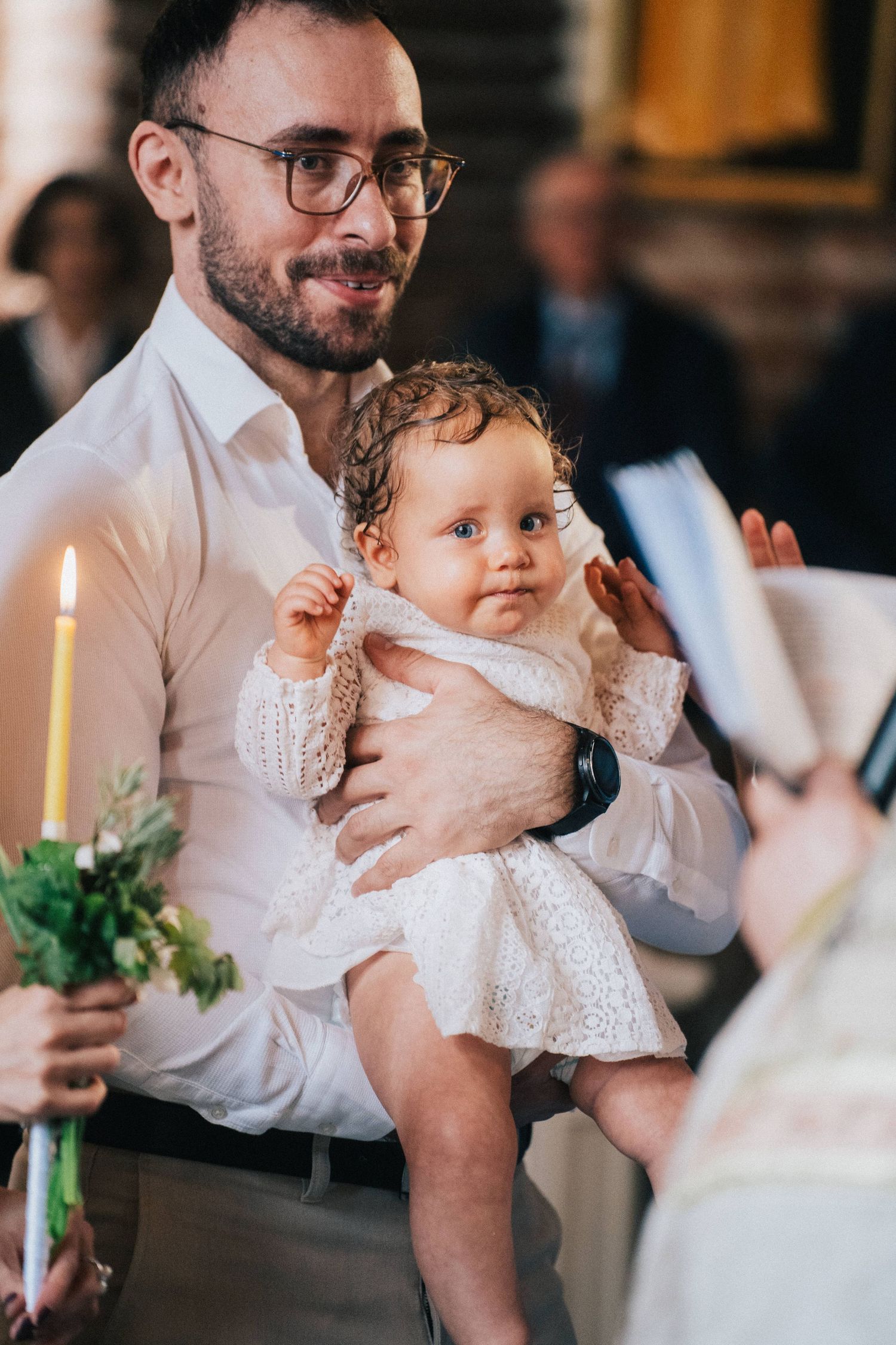A bearded father with glasses holds his baby girl in a white lace dress, while a lit candle burns during the church baptism.