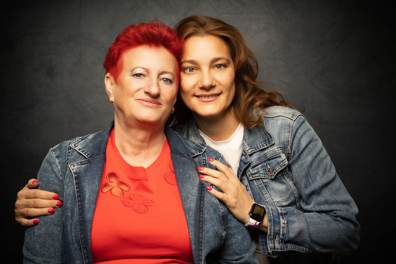 A red-haired woman in denim jacket and a young woman in a matching jacket embrace in a warm studio portrait.