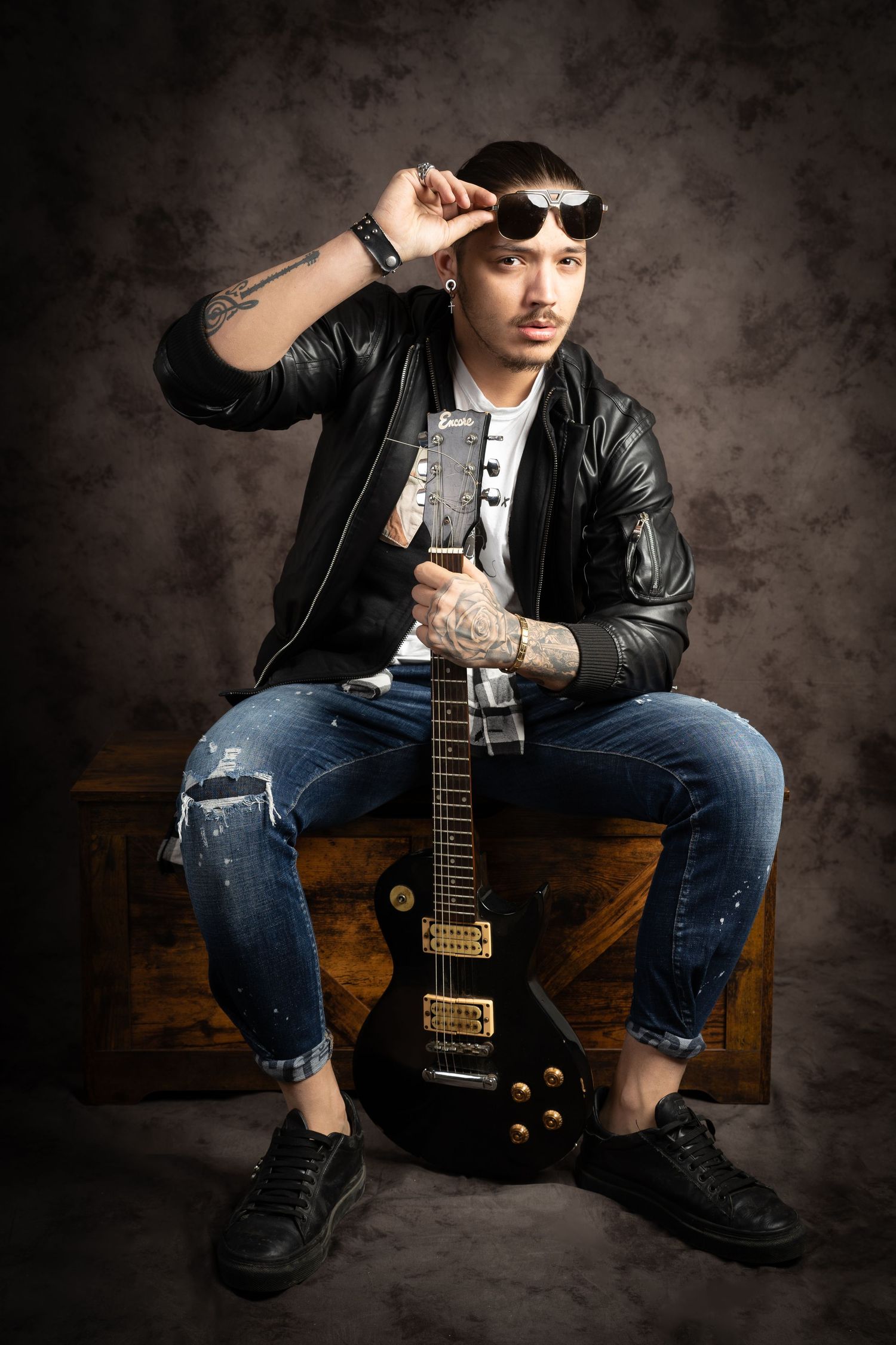 Tattooed man in sunglasses and a black leather jacket sits on a wooden trunk holding an electric guitar in a studio portrait.