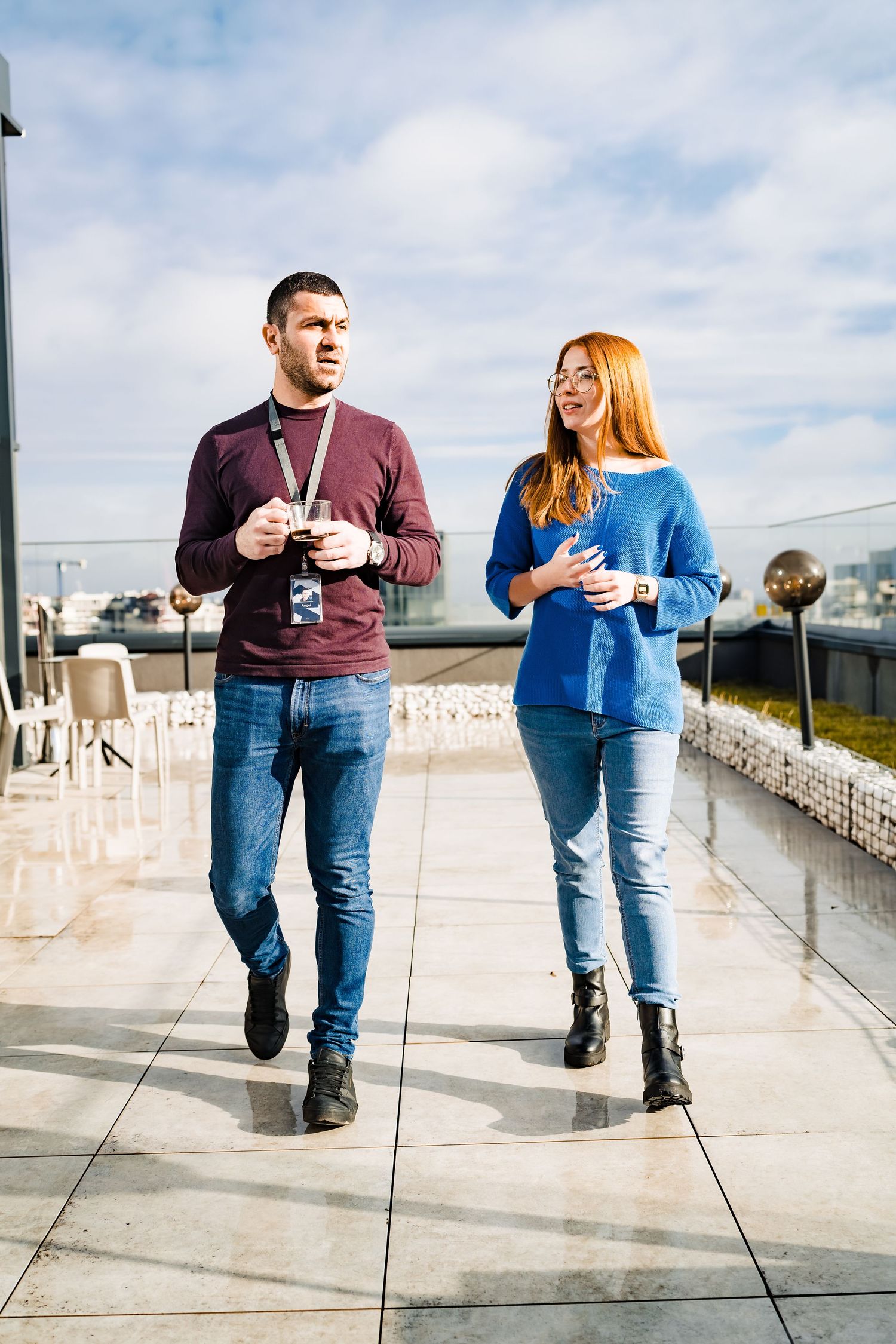 A man with a lanyard and a woman in a blue sweater chat while walking across a bright rooftop terrace.